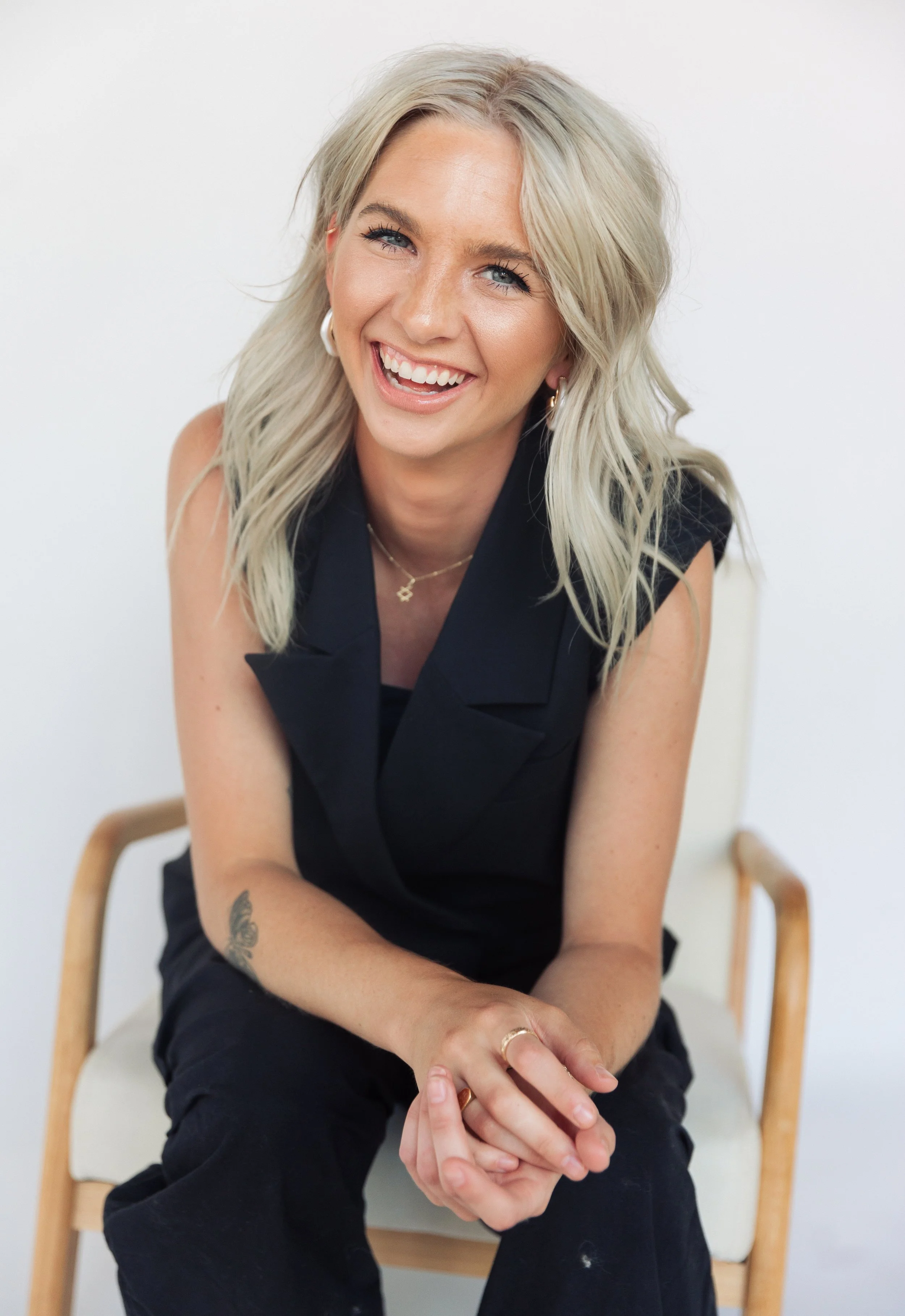A smiling woman with blonde hair, wearing a black sleeveless top, sitting on a beige chair with wooden armrests, against a plain white background.