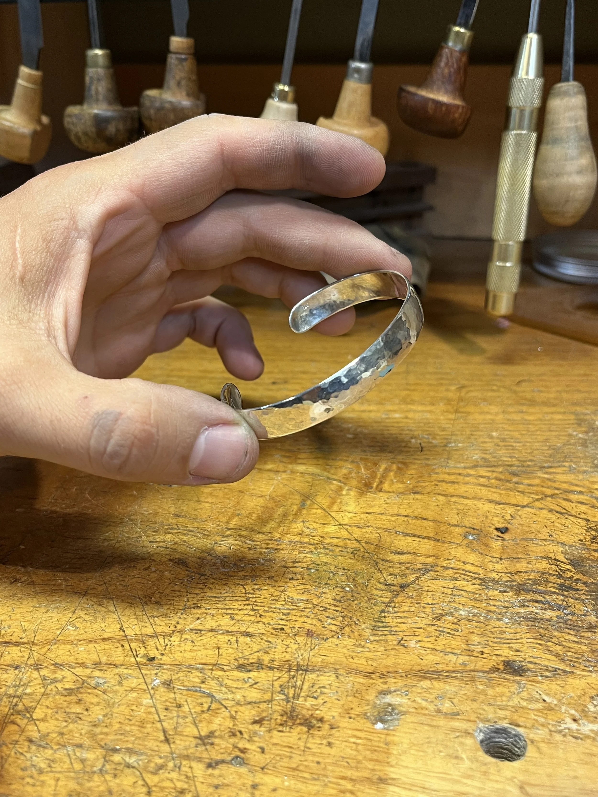 A person holding a hammered silver cuff bracelet with a textured finish, with woodworking tools in the background on a wooden workbench.
