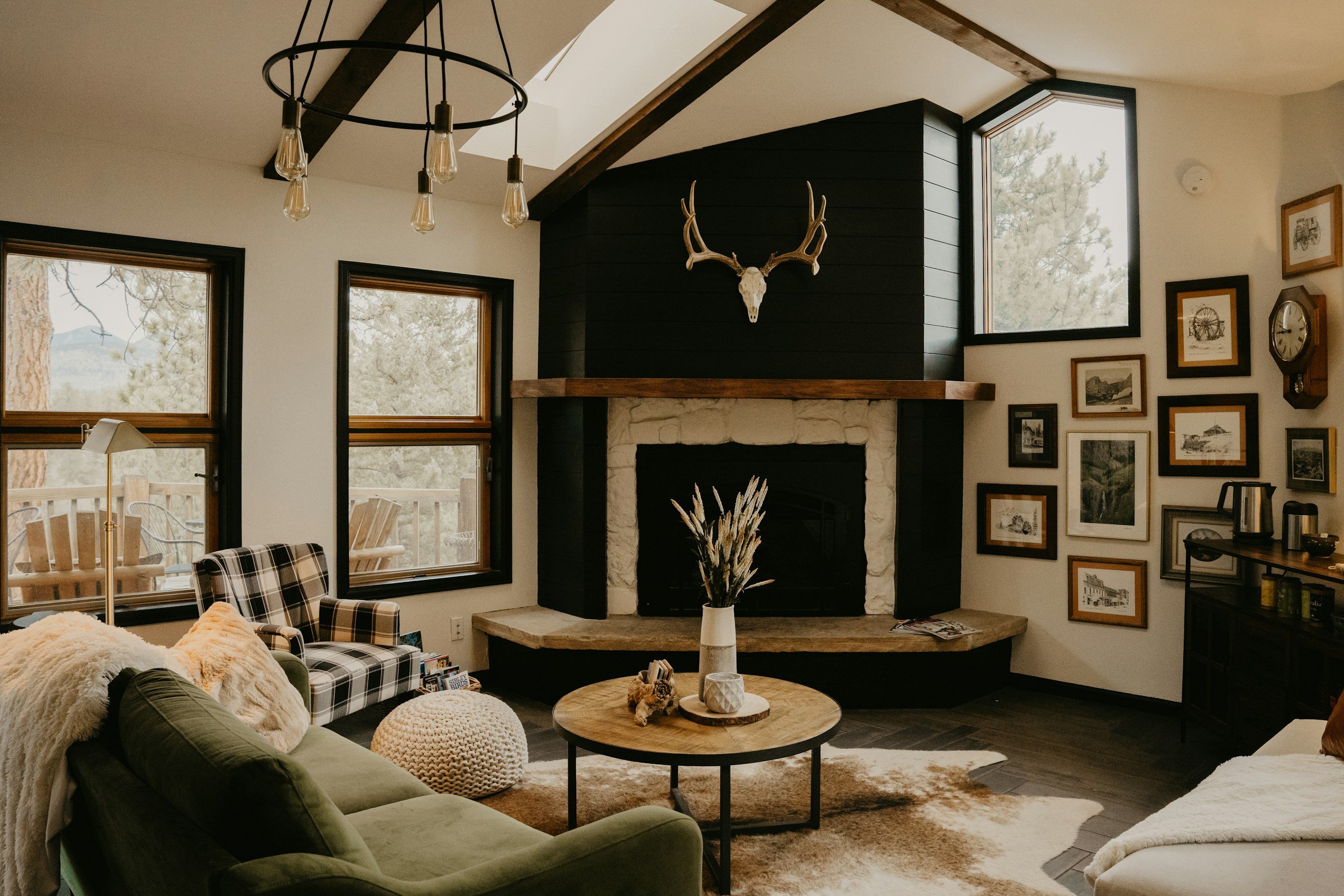 Cozy living room with a stone fireplace, mounted deer antlers, and framed artwork on the wall. There are large windows, a plaid armchair, a green sofa with pillows, a round wooden coffee table with decorative items, and a cowhide rug.