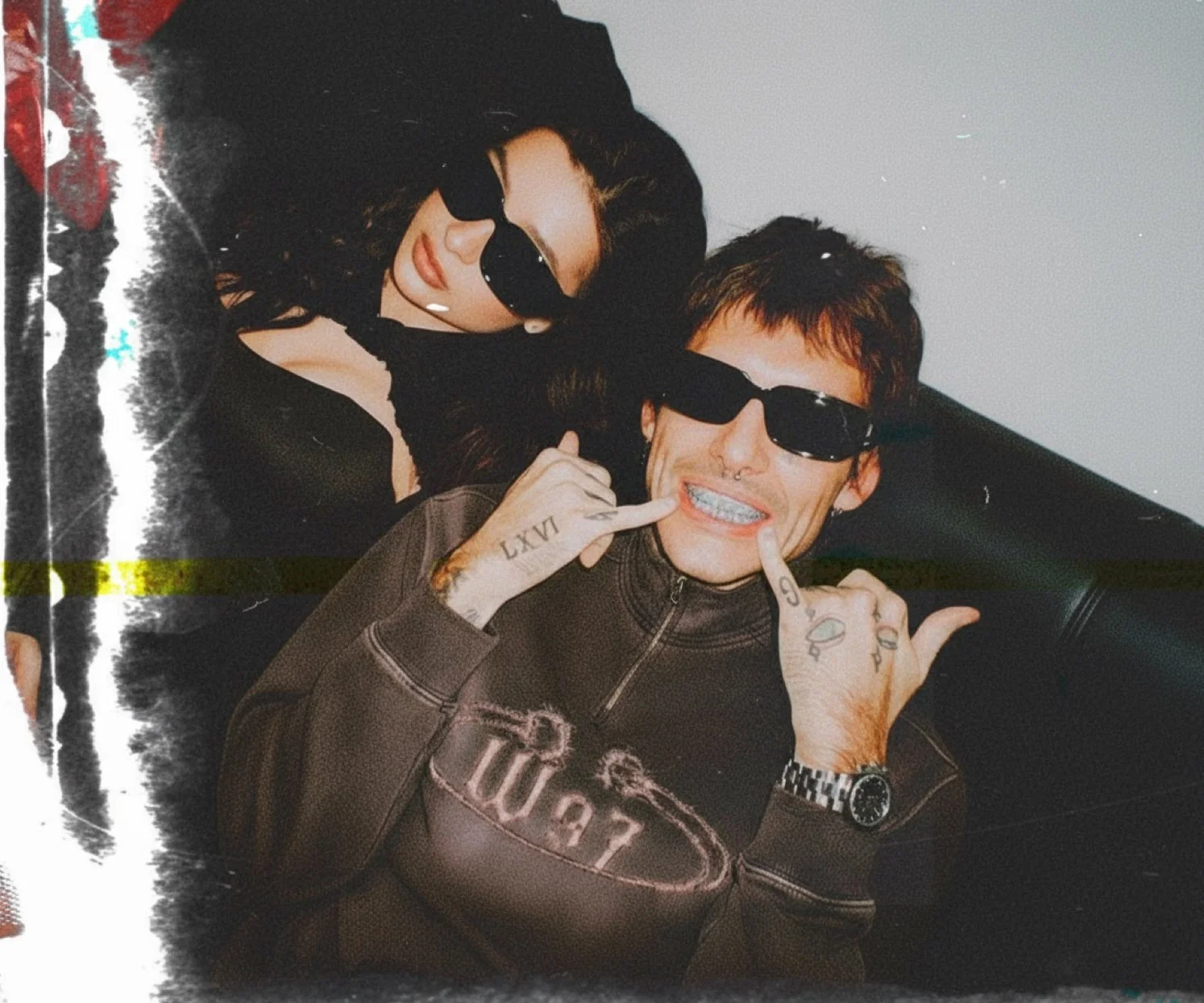Two people, a woman with long dark hair and sunglasses, and a man with short hair, tattoos, and sunglasses, are sitting close together on a black leather couch, both making a gesture with their fingers near their faces.