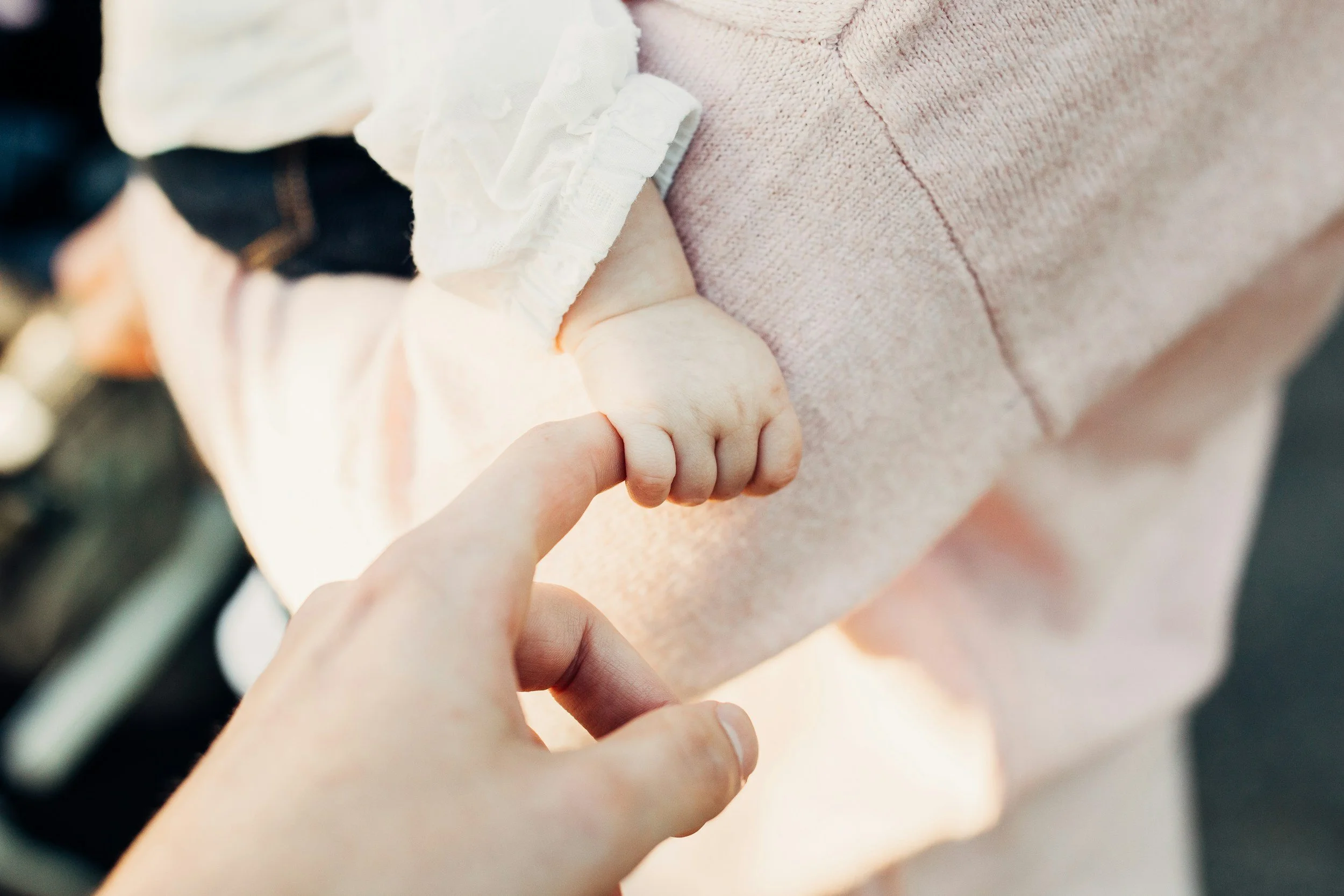 A close-up of an adult's finger gently holding a baby's tiny hand, with the baby wearing a pink shirt and a white sleeve.