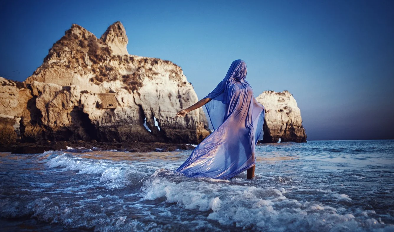 A woman in a flowing blue dress swimming in the ocean near rocky cliffs during twilight.