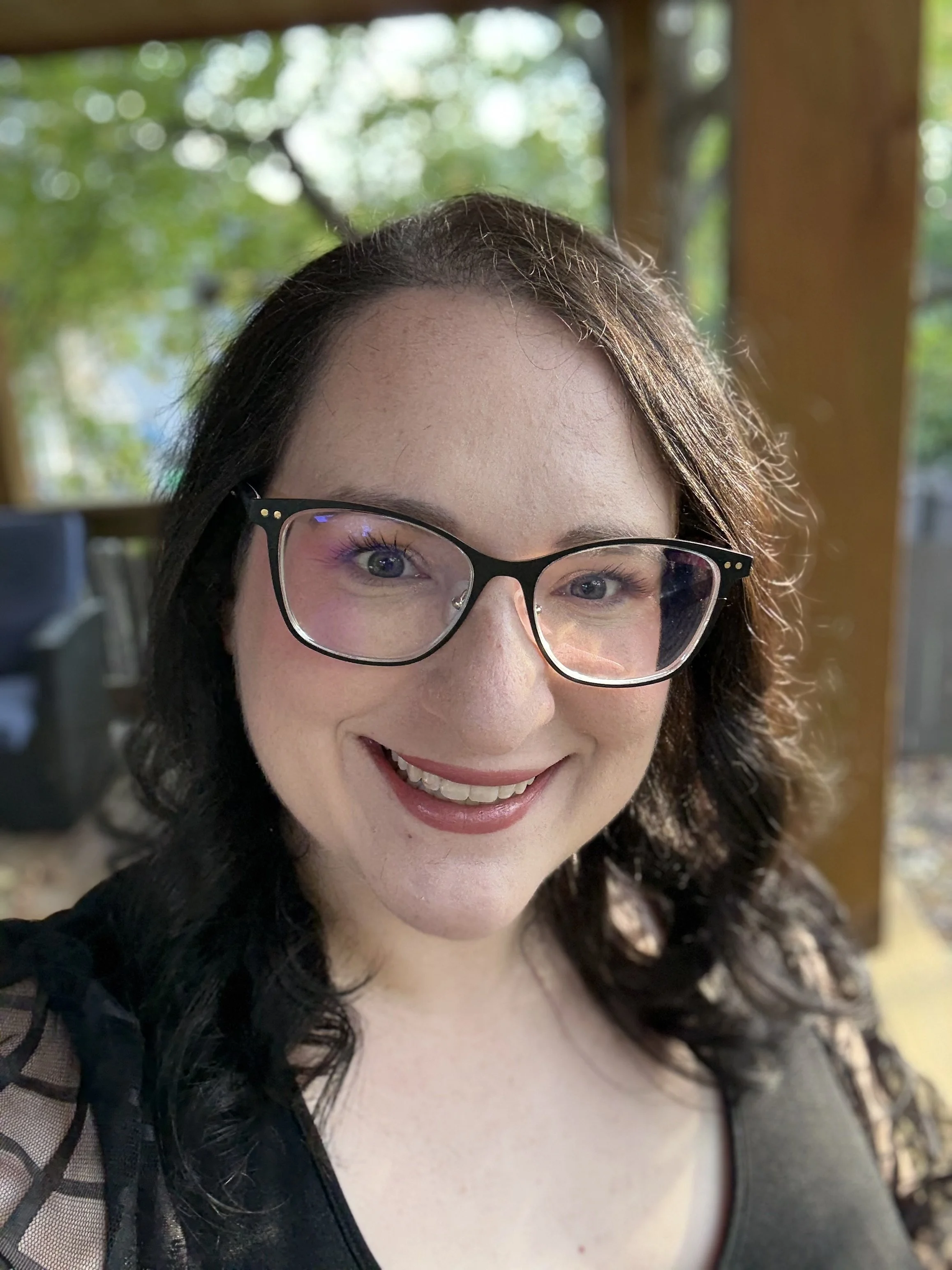 A woman with curly dark hair, wearing glasses, smiling outdoors with a green background.