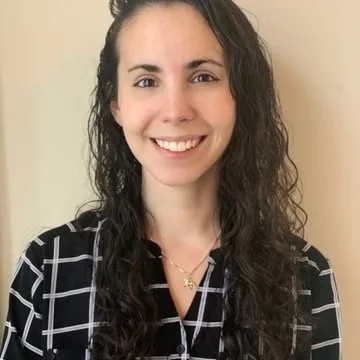 A woman with long, curly dark hair smiling at the camera, wearing a black and white checkered shirt and a gold necklace, standing against a beige wall.