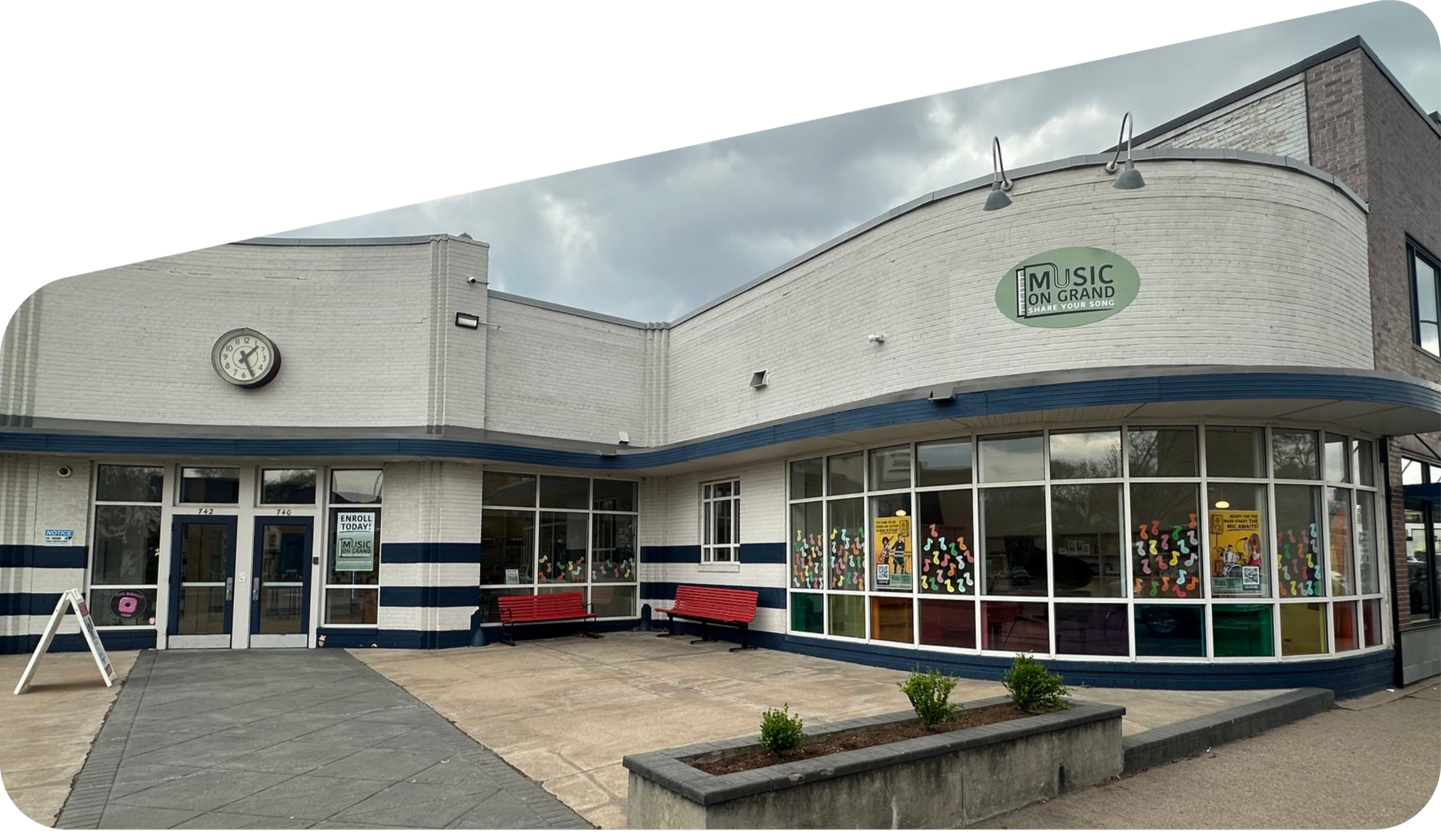 The exterior of a music school called 'Music on Grand' with a clock showing 3:11. The building has a white brick facade with large windows decorated with colorful musical note stickers. There are red benches outside and a small landscaped area with shrubs. The sky is overcast.