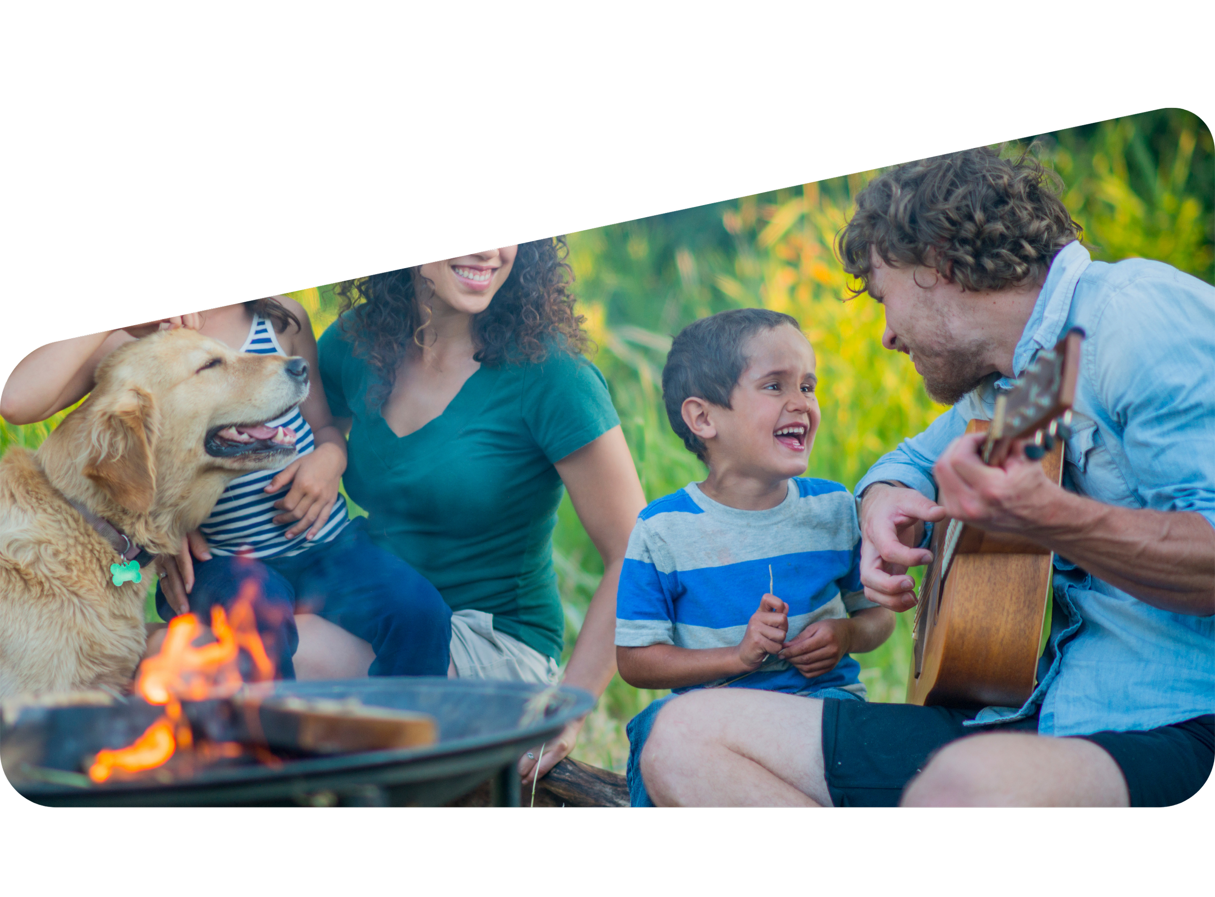 A group of people, including children and adults, gathered outdoors around a campfire during daytime. One person is playing a guitar, and there is a dog among them. The background features green trees and foliage.