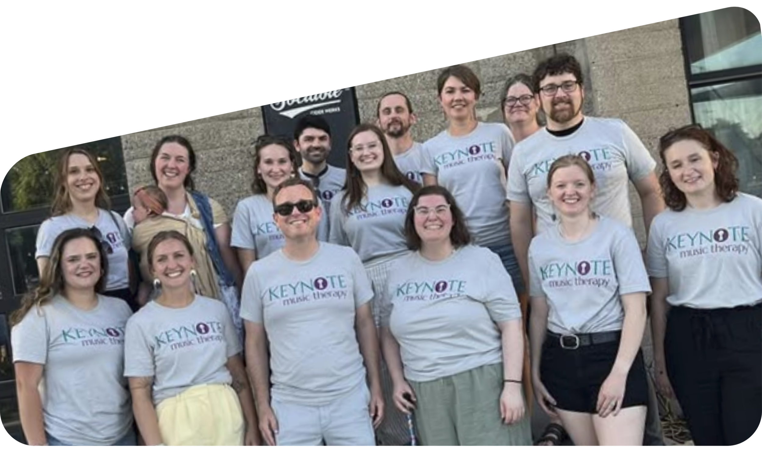 Group of smiling people wearing white T-shirts with 'Keynote Music Therapy' logo, standing outside in front of a brick building with a sign. It appears to be a sunny day.