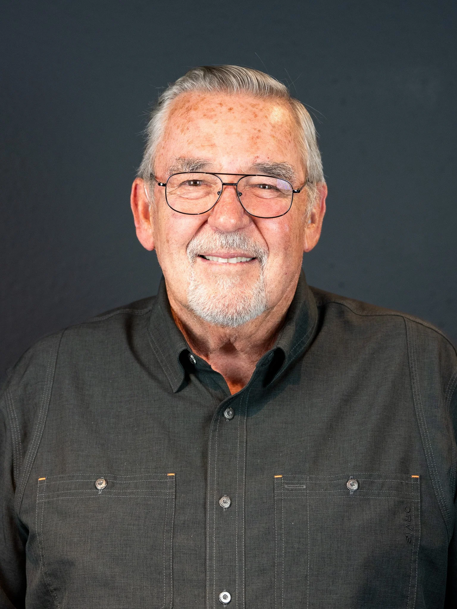 An elderly man with gray hair, glasses, and a beard wearing a dark button-up shirt, smiling in front of a dark background.