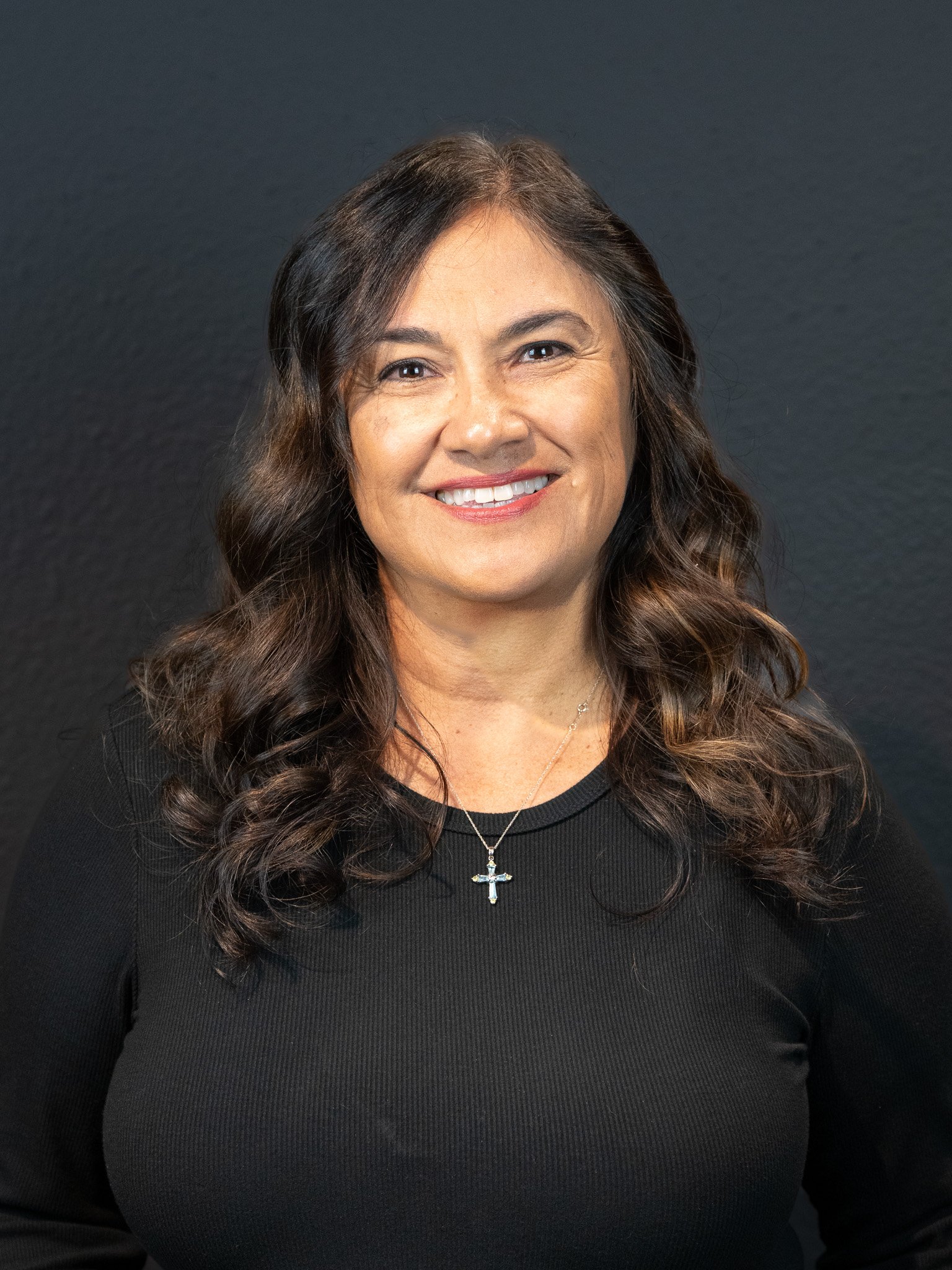 A woman with long, wavy brown hair smiling, wearing a black top and a cross necklace, against a dark background.