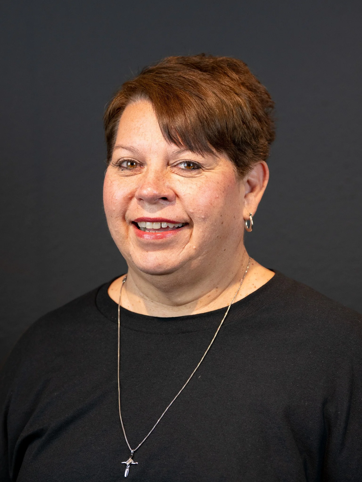 Portrait of a middle-aged woman with short brown hair, smiling, wearing a black top and a silver necklace with a cross pendant, against a dark background.