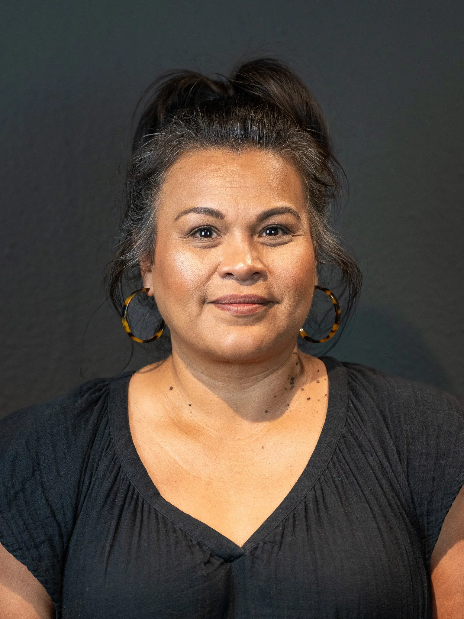 A woman with dark hair styled in a high ponytail, wearing large hoop earrings and a black top, standing against a dark background.