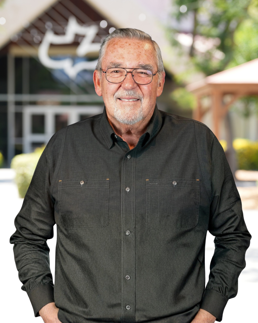 An elderly man with gray hair, glasses, and a beard wearing a dark button-up shirt, smiling in front of a dark background.