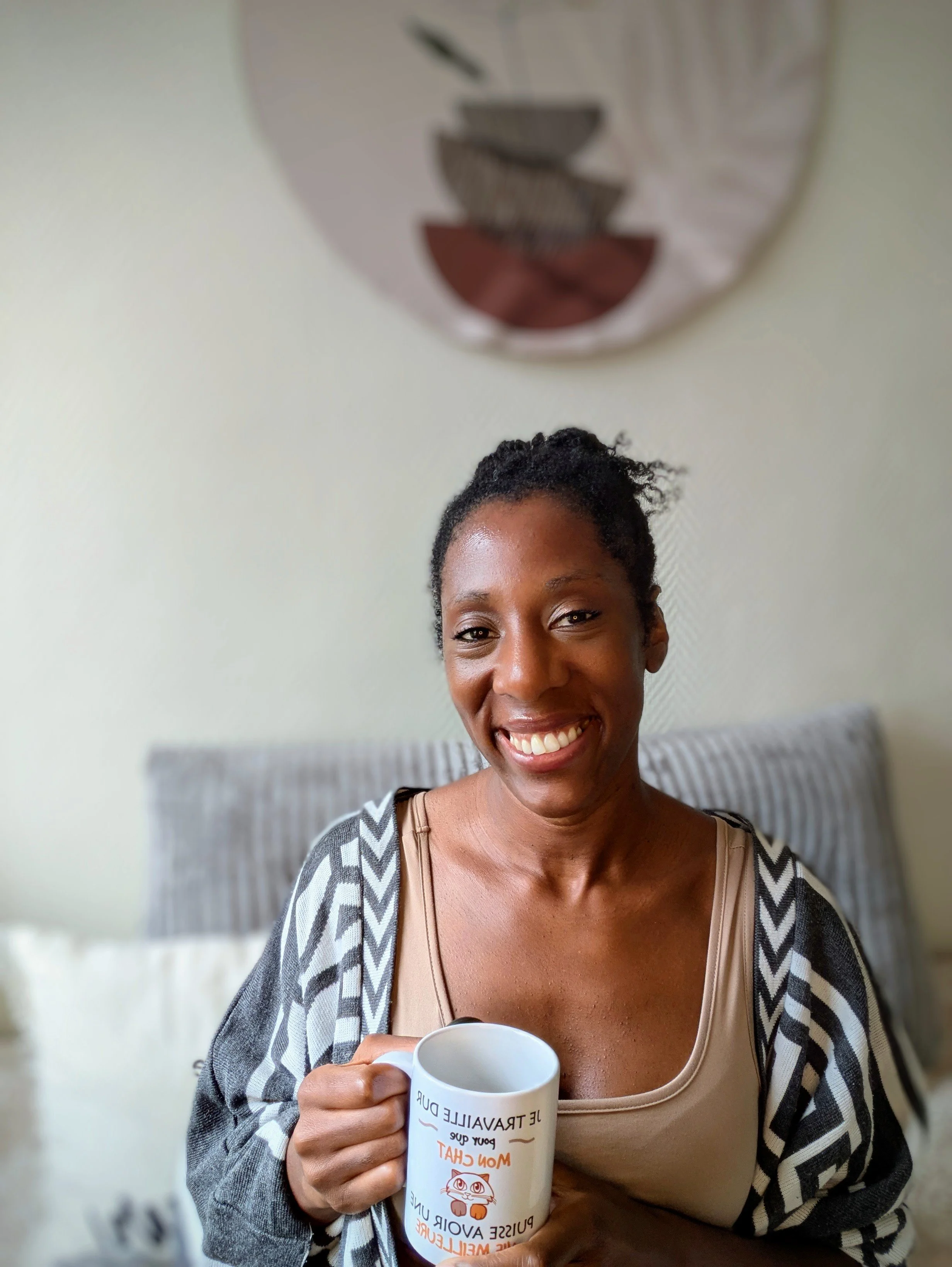 Une femme souriante tenant une tasse blanche dans une pièce. En arrière-plan, un lit avec une tête de lit rayée et un tableau mural abstrait.