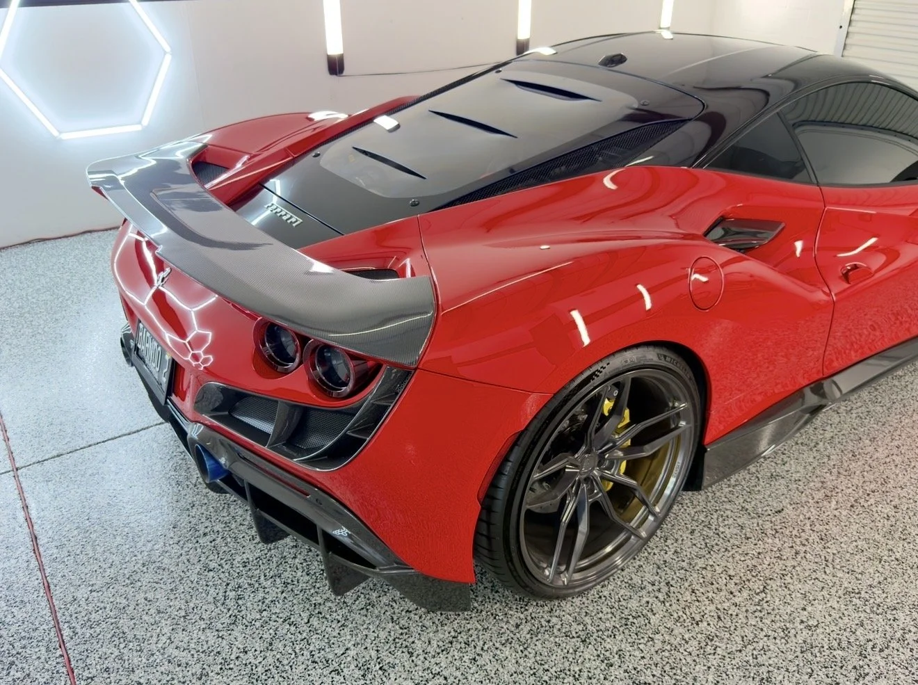 Red and black Ferrari sports cars parked indoors, with a carbon fiber rear wing on the red car and a hexagonal neon light on the wall.