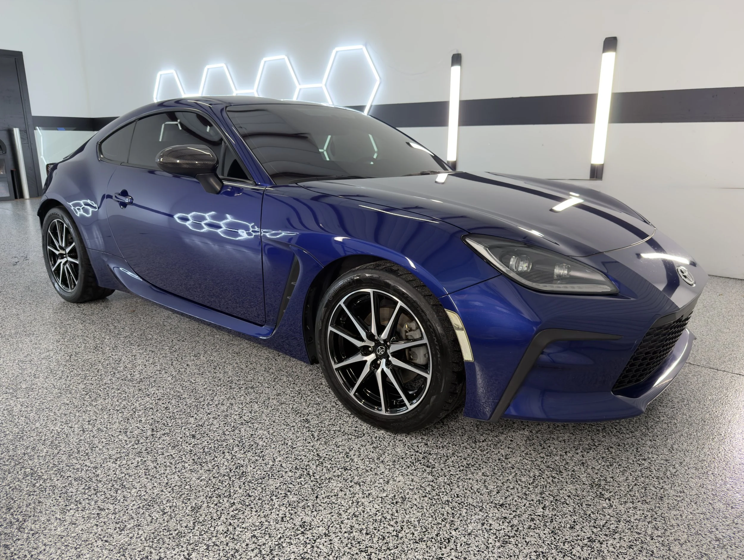 A blue sports car with black wheels parked indoors against a white wall with black accents and modern lighting.