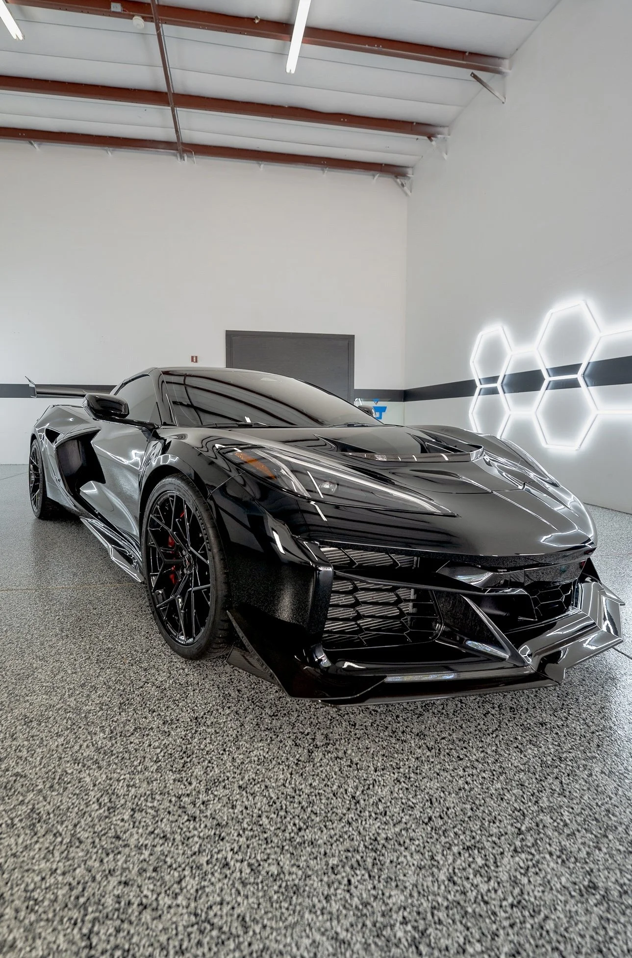 A sleek black sports car with sharp lines and tinted windows parked indoors against a white wall with hexagonal light fixtures.