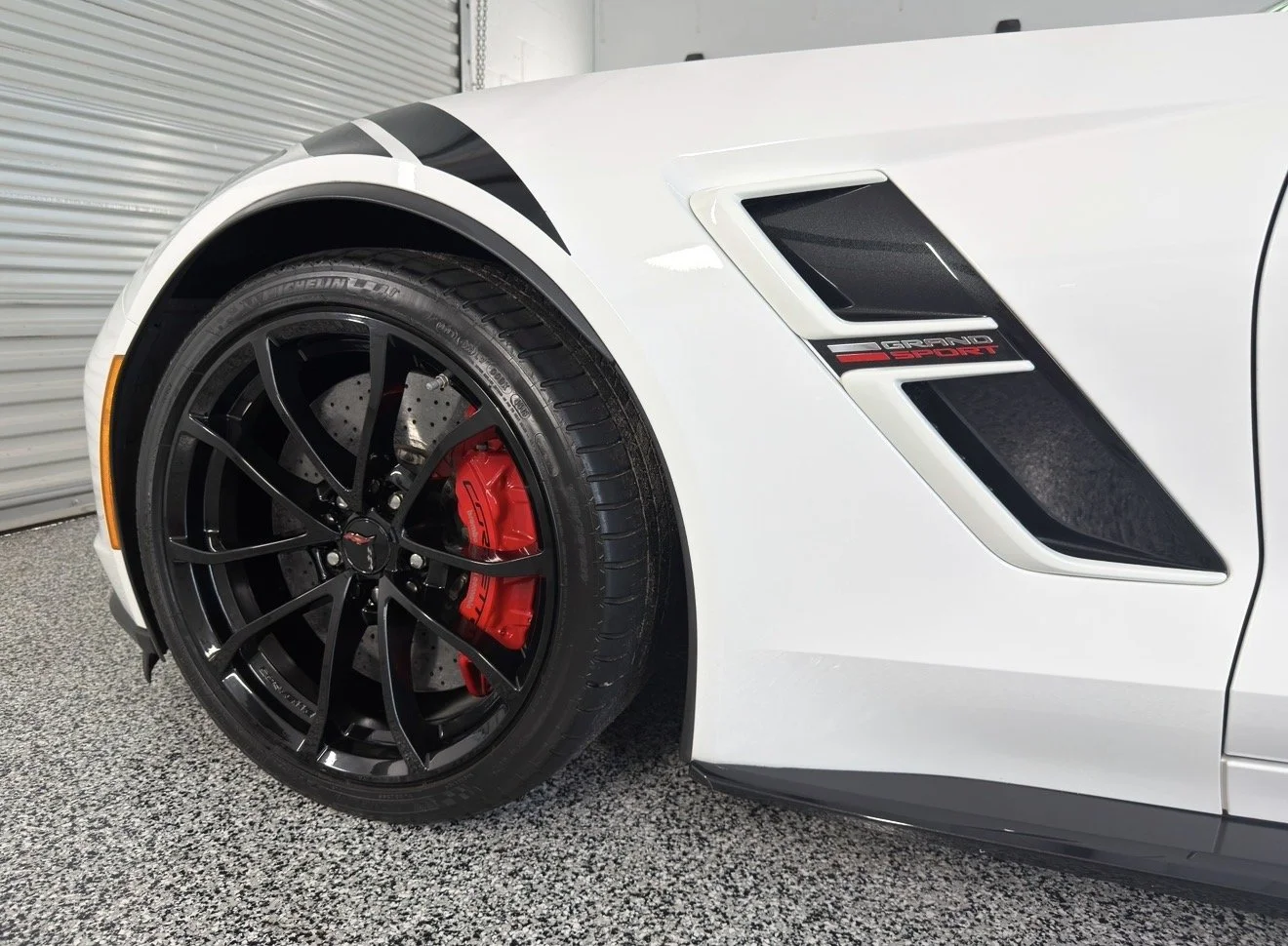 Close-up of a white Chevrolet Corvette Grand Sport car's front left side, showcasing a black wheel with red brake caliper, and black side vent with 'Grand Sport' emblem.