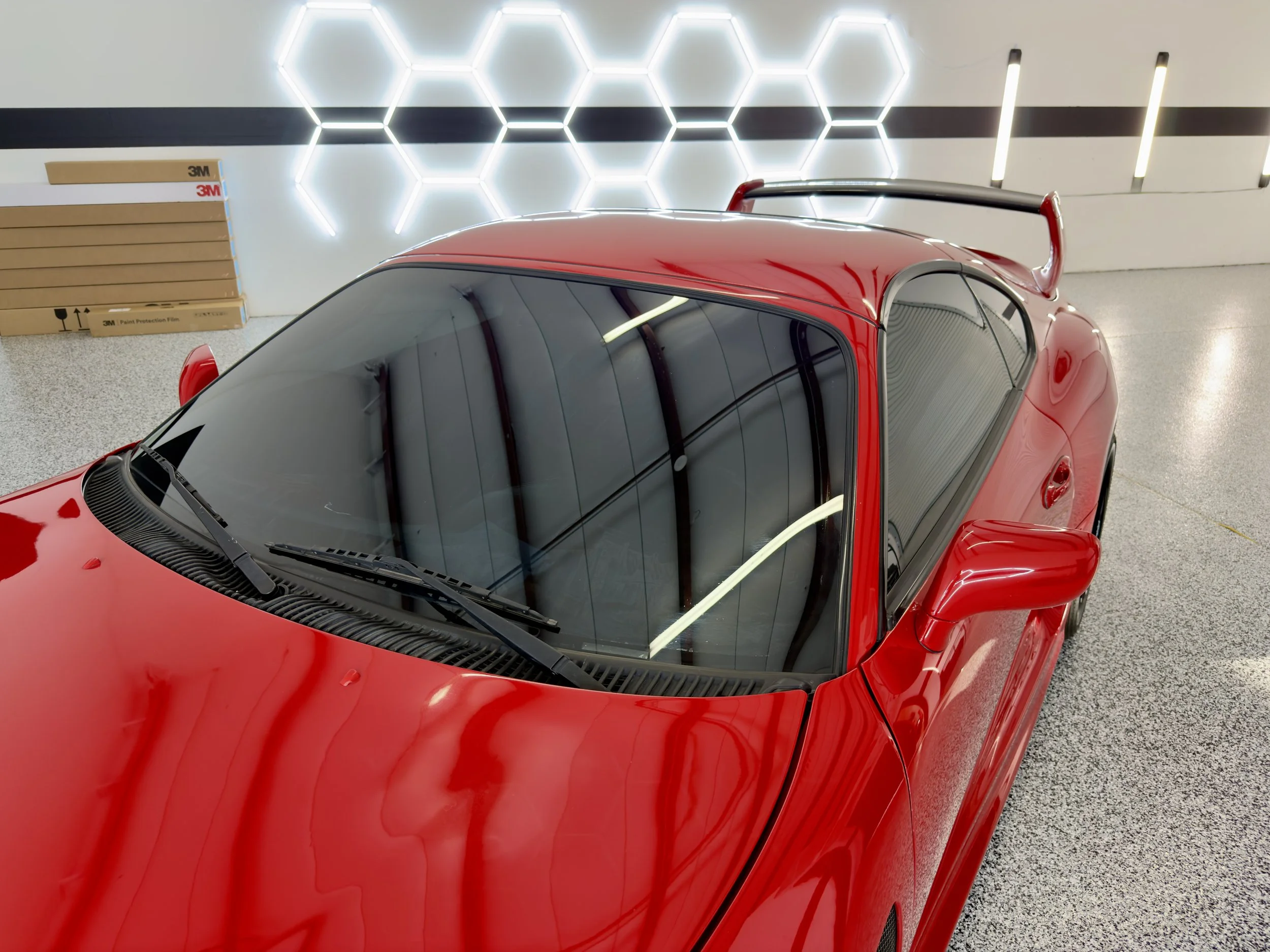 A red sports car with a black and red spoiler parked indoors, with a white wall featuring illuminated hexagon designs and a stack of cardboard boxes labeled "3M" in the background.
