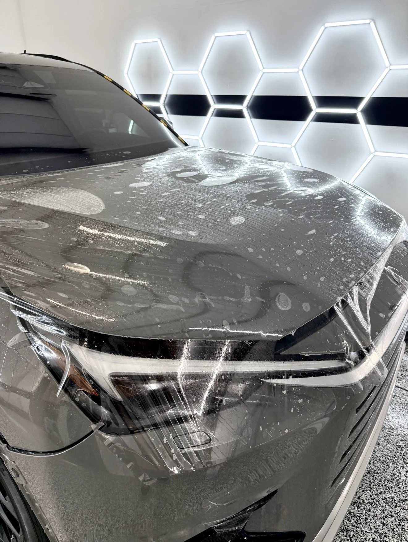 A silver car being wrapped in a clear protective film in a modern garage with illuminated hexagonal wall patterns.