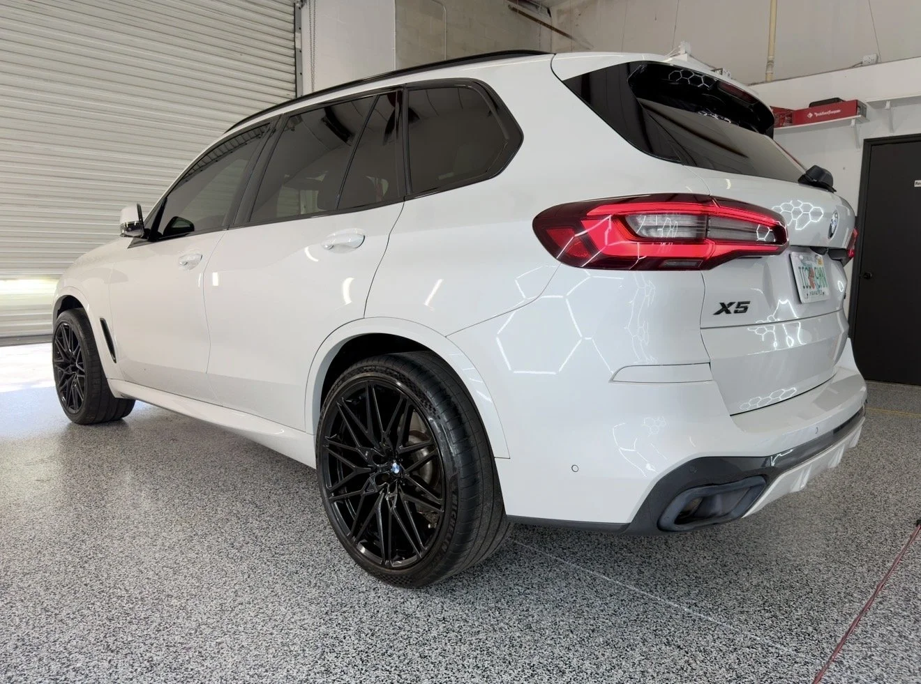 White BMW X5 parked indoors with black wheels, rear taillights, and a honeycomb pattern on the rear window.