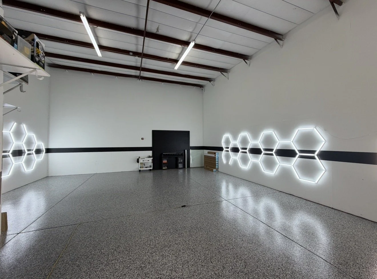 Empty room with white walls, illuminated hexagon-shaped light fixtures on the side walls, a black stripe running horizontally around the room, and a grey concrete floor. There are some items on the left and right sides near the back corner.