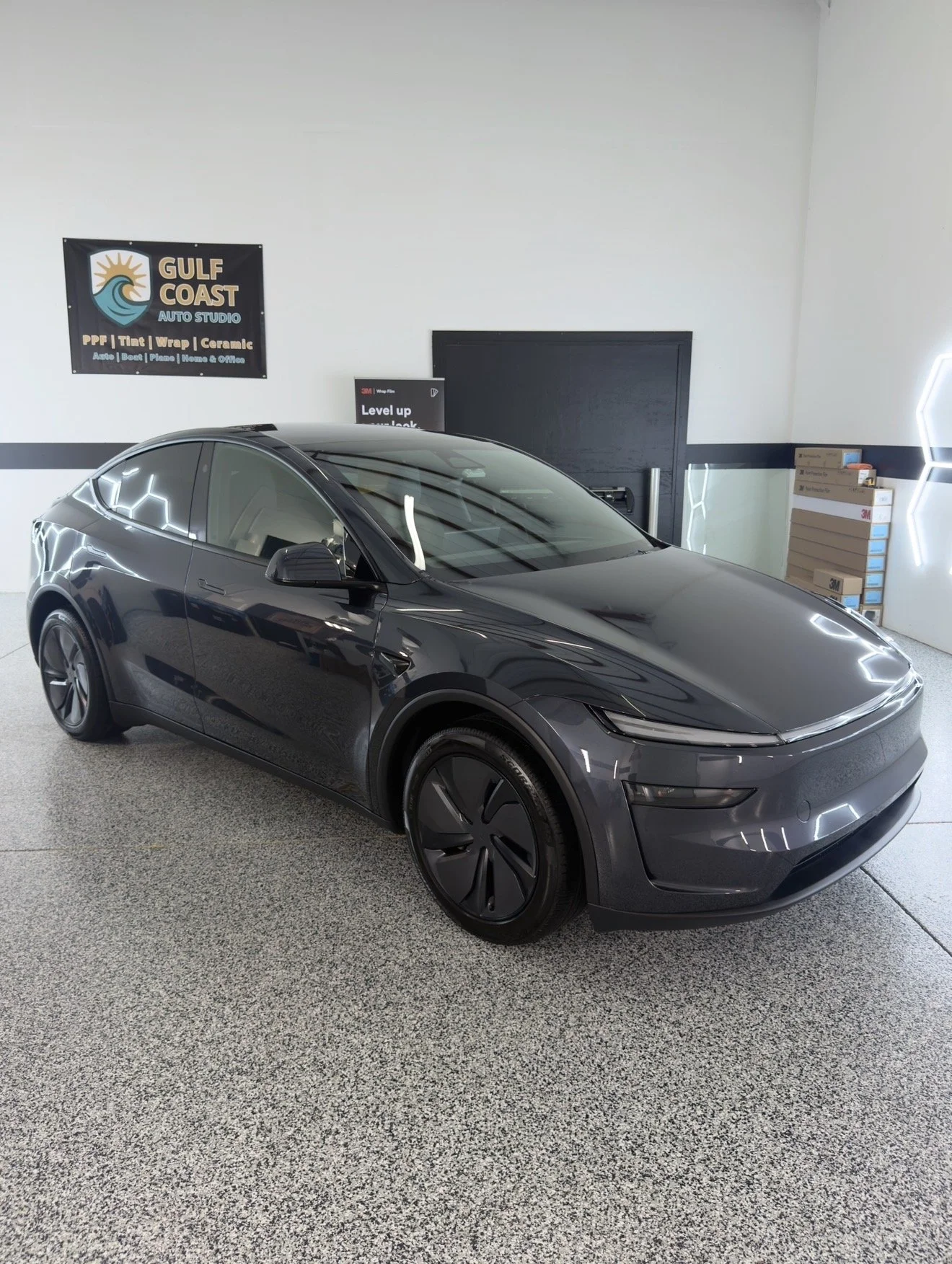 A black Tesla Model Y parked indoors at Gulf Coast Auto Studio, with a wall sign advertising vehicle detailing services and some boxes of 3M products in the background.