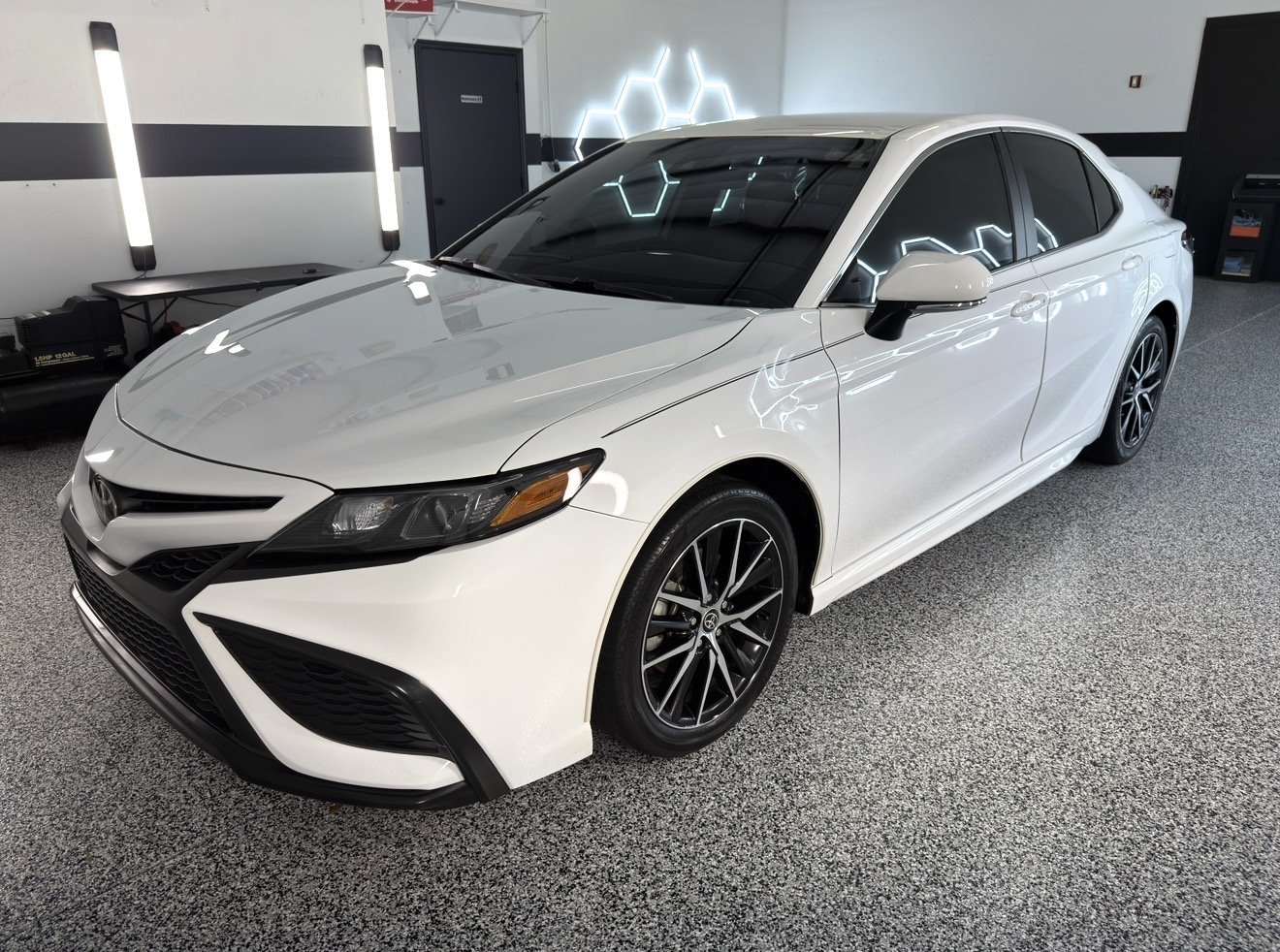 A white sedan car parked indoors, with black rims and tinted windows, inside a modern garage or showroom with gray flooring and LED hexagon wall lights.