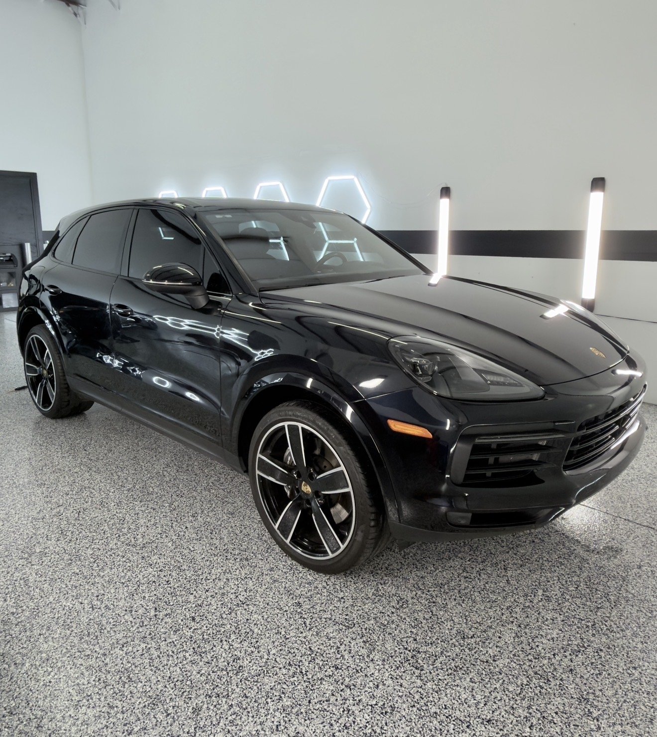 A black Porsche Macan SUV parked indoors with a plain wall behind, illuminated by vertical and hexagonal LED lights.