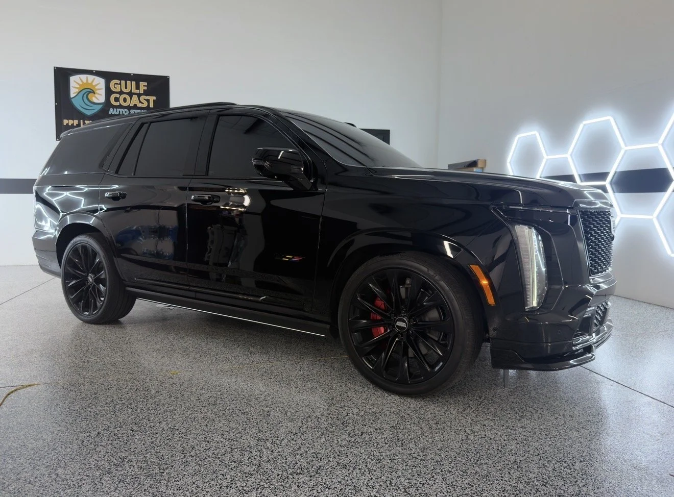 Black luxury SUV with tinted windows parked indoors, illuminated hexagonal wall lights, and a Gulf Coast Auto Spa sign in the background.