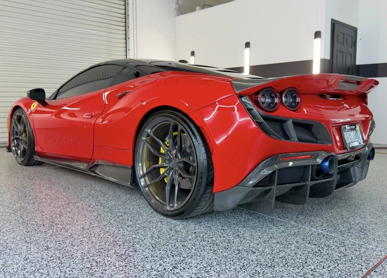 Red race car with black accents parked indoors on speckled floor, featuring aerodynamic design, large rear wing, black wheel rims, yellow brake calipers, and blue exhaust tips.