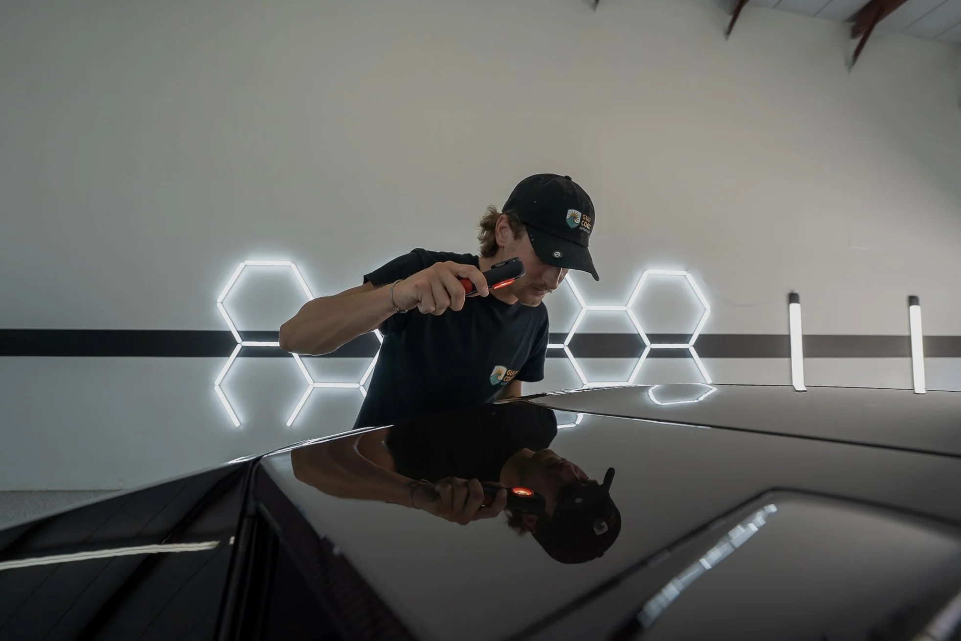 A person wearing a black Cap with a logo and black shirt while inspecting a shiny black car in a modern room with hexagonal light fixtures on the wall.