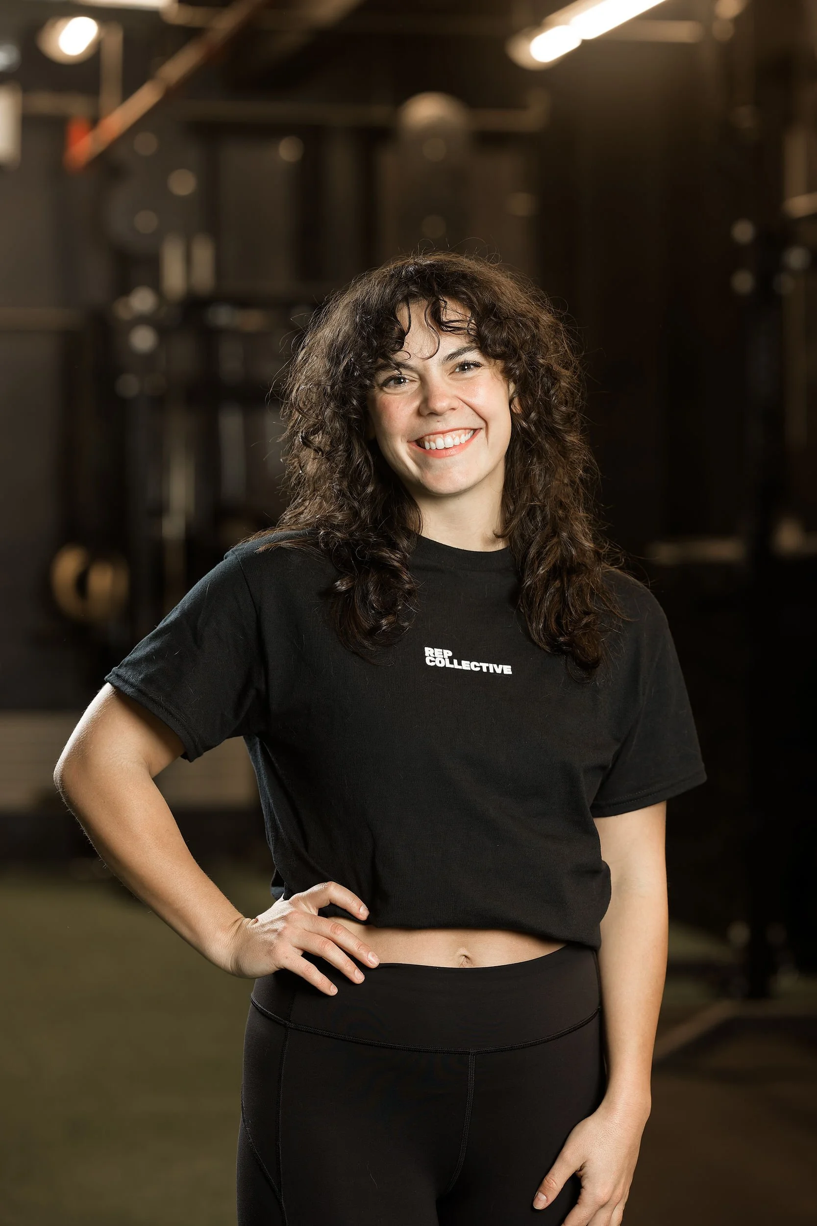 A woman with curly dark hair smiling, wearing a black crop top and black leggings, in a gym setting.
