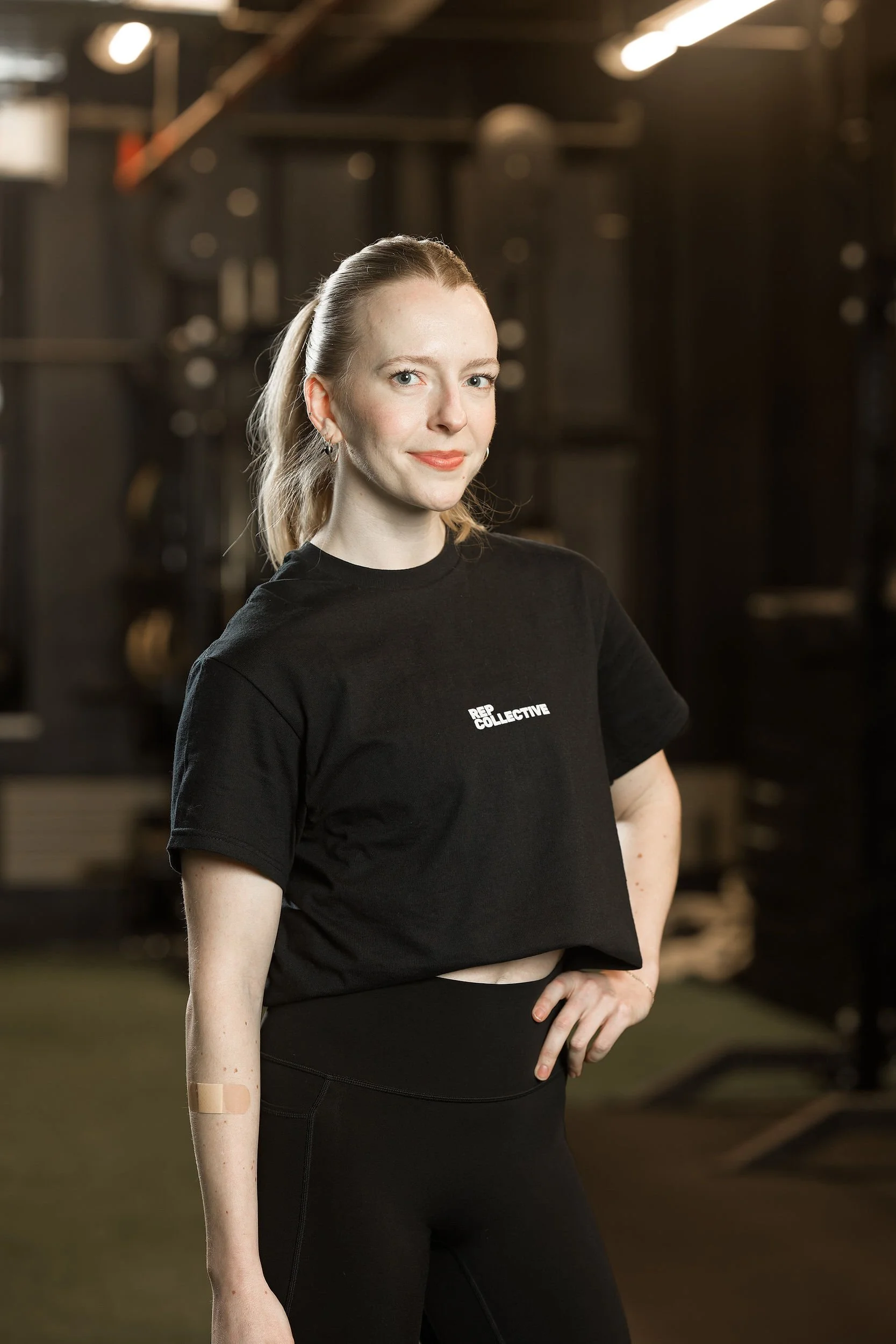 A woman standing in a gym, wearing a black t-shirt with 'REPLICA COLLECTIVE' written on it and black leggings. She has blonde hair tied back and is smiling softly. The background features gym equipment and a dimly lit environment.