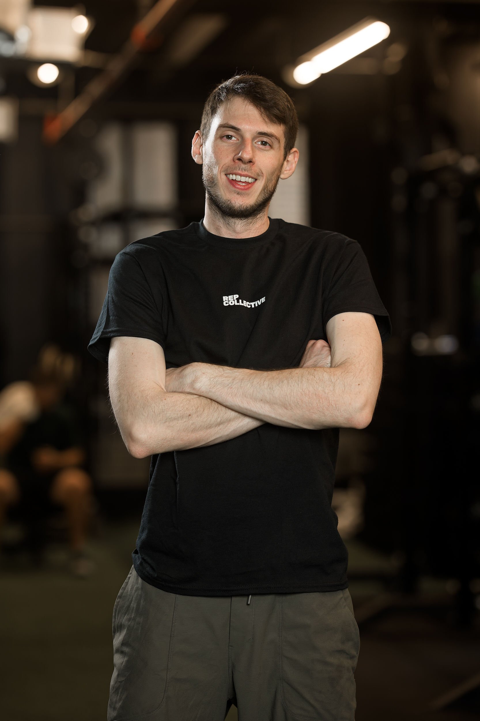 A young man with short brown hair and light skin, smiling and crossing his arms, standing in a gym or fitness center.
