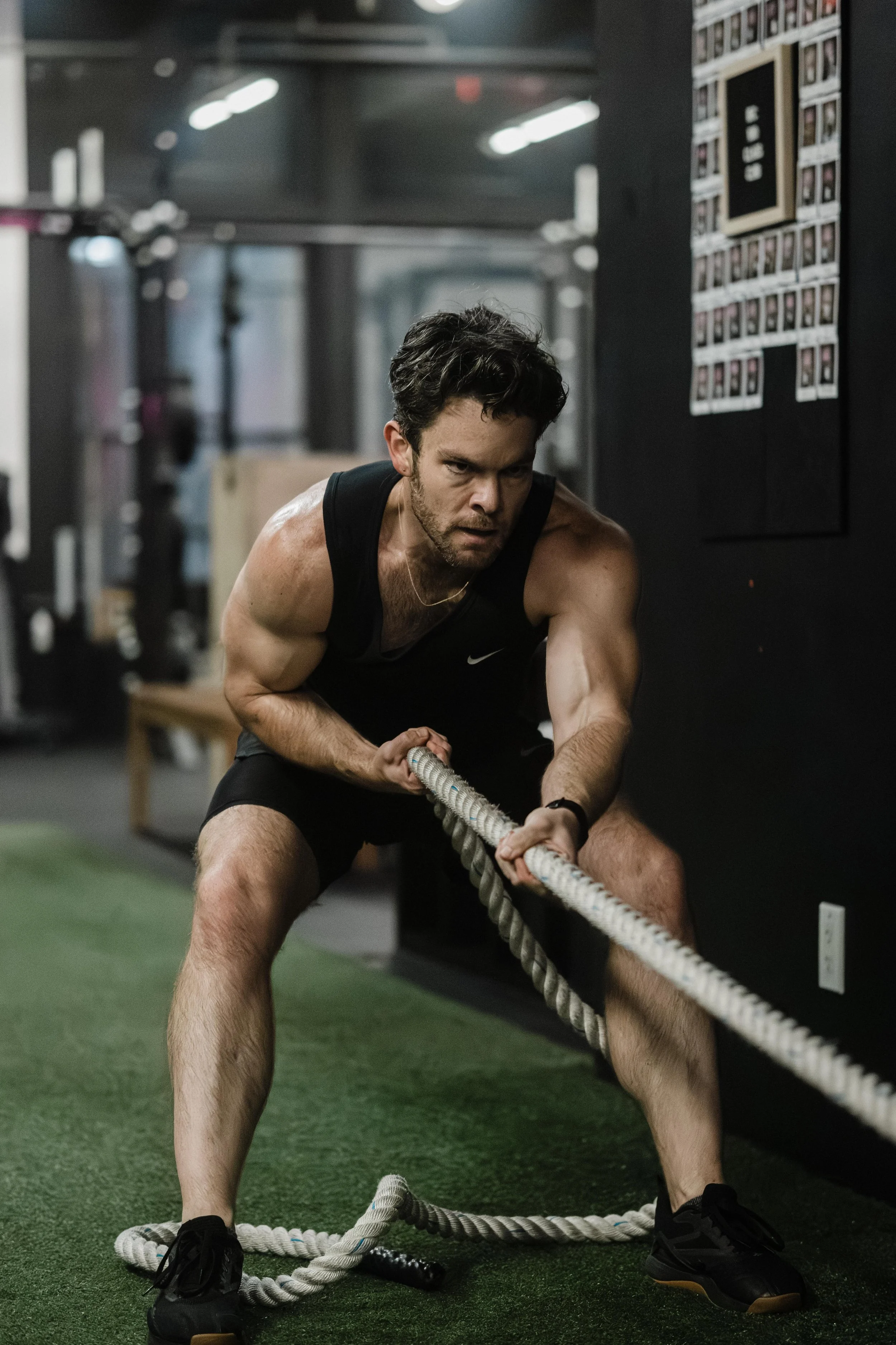 A man with dark hair, wearing a black sleeveless athletic shirt and shorts, is intensely pulling on a battle rope during a workout at a gym.