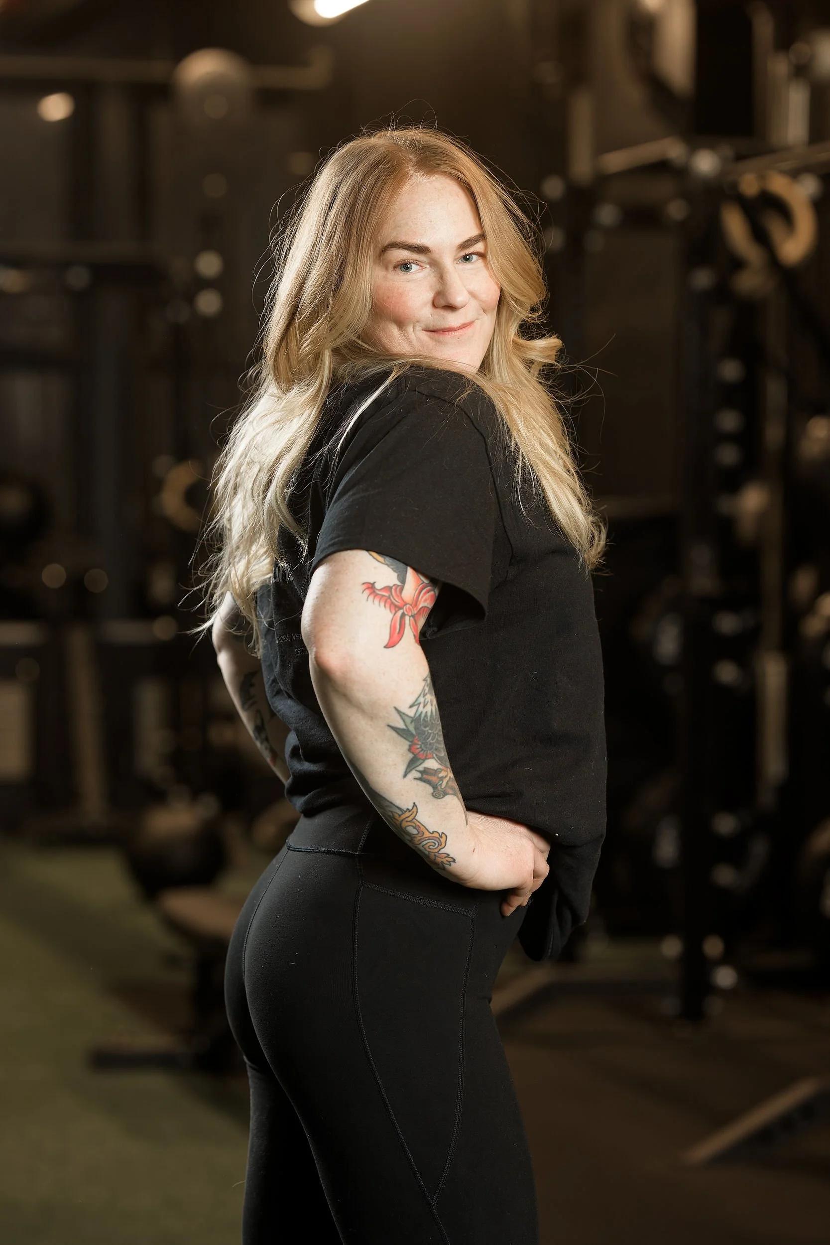 A woman with long blonde hair and tattoos on her left arm, wearing a black shirt and black pants, standing in a gym. She is smiling and has her hands on her hips.