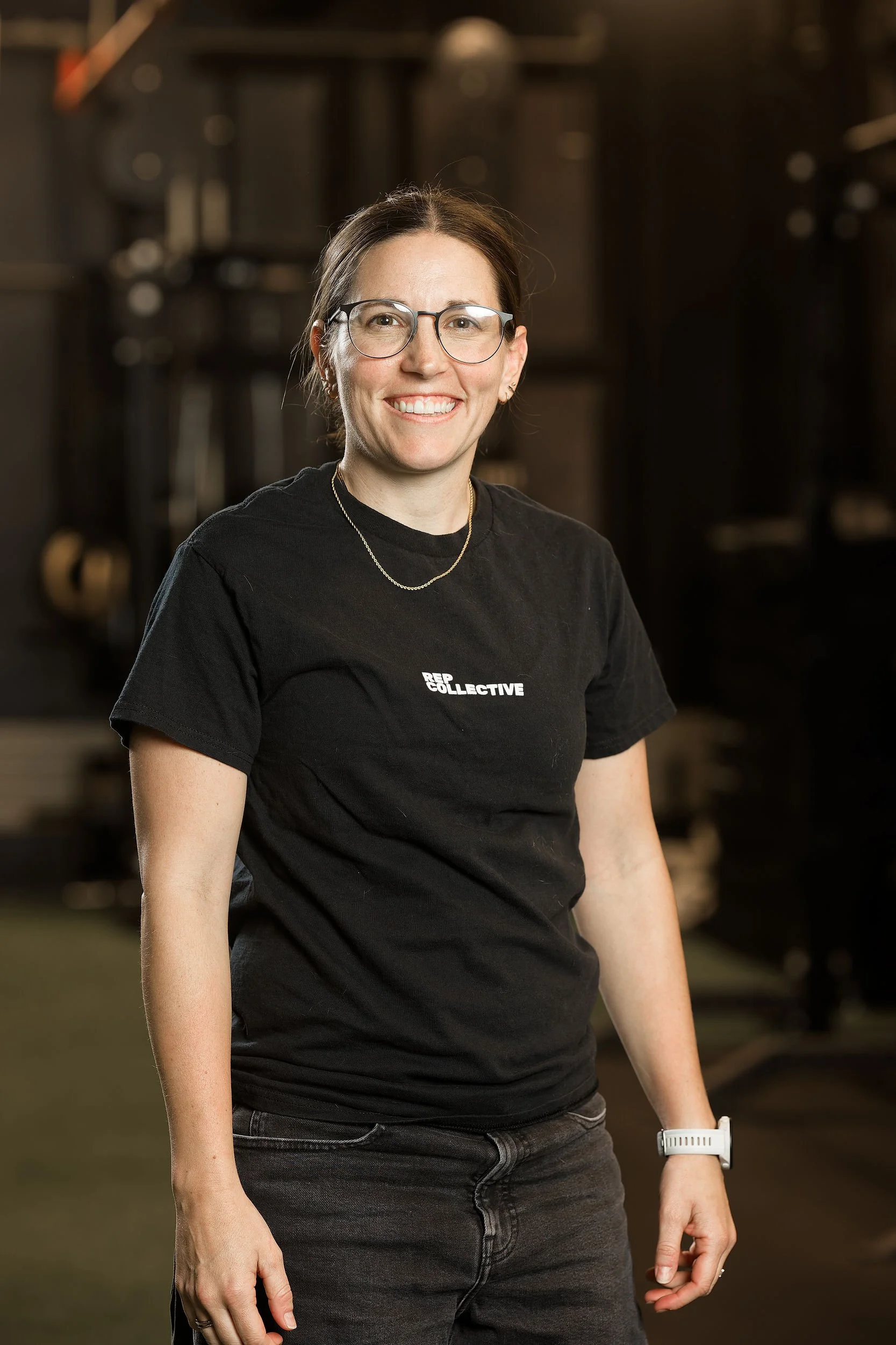 A smiling woman with glasses and brown hair, wearing a black 'REPS COLLECTIVE' t-shirt and a white wristwatch, standing in a gym or fitness studio.