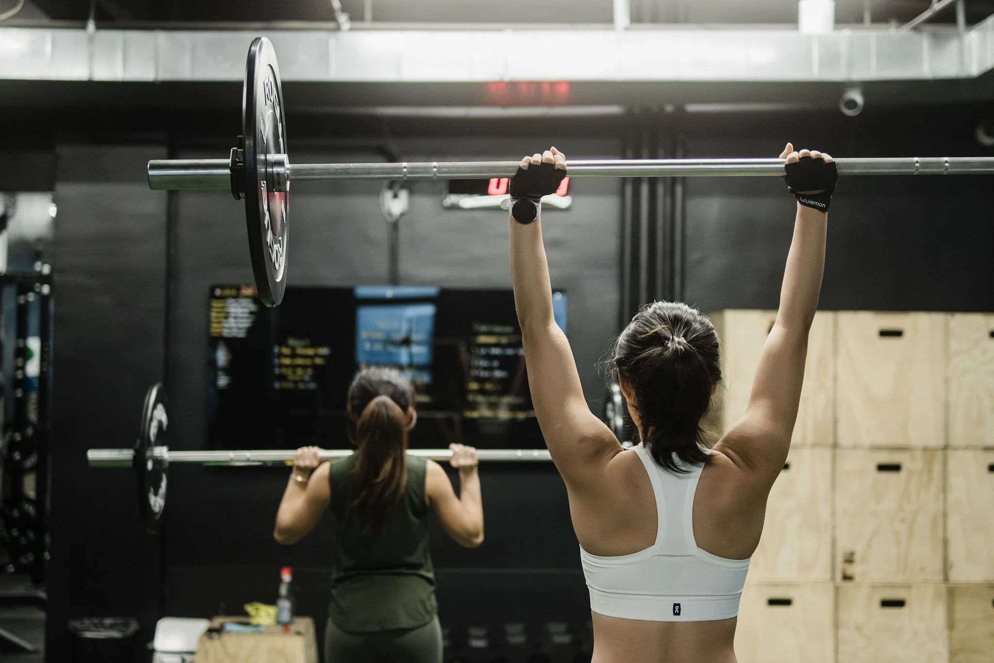 Two women lifting barbells during a workout in a gym, with one woman in the foreground wearing a white sports bra and the woman in the background wearing a dark tank top