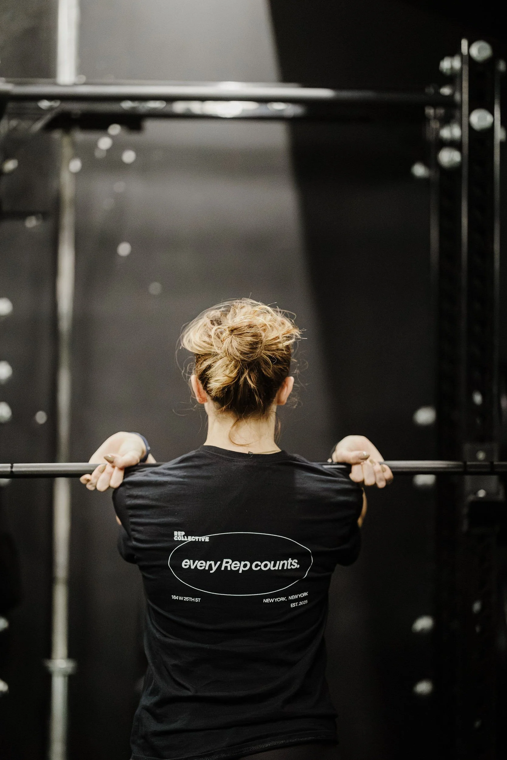 A woman with styled hair, wearing a black t-shirt with the phrase 'every Rep counts.' printed on the back, is lifting a barbell in a gym setting.