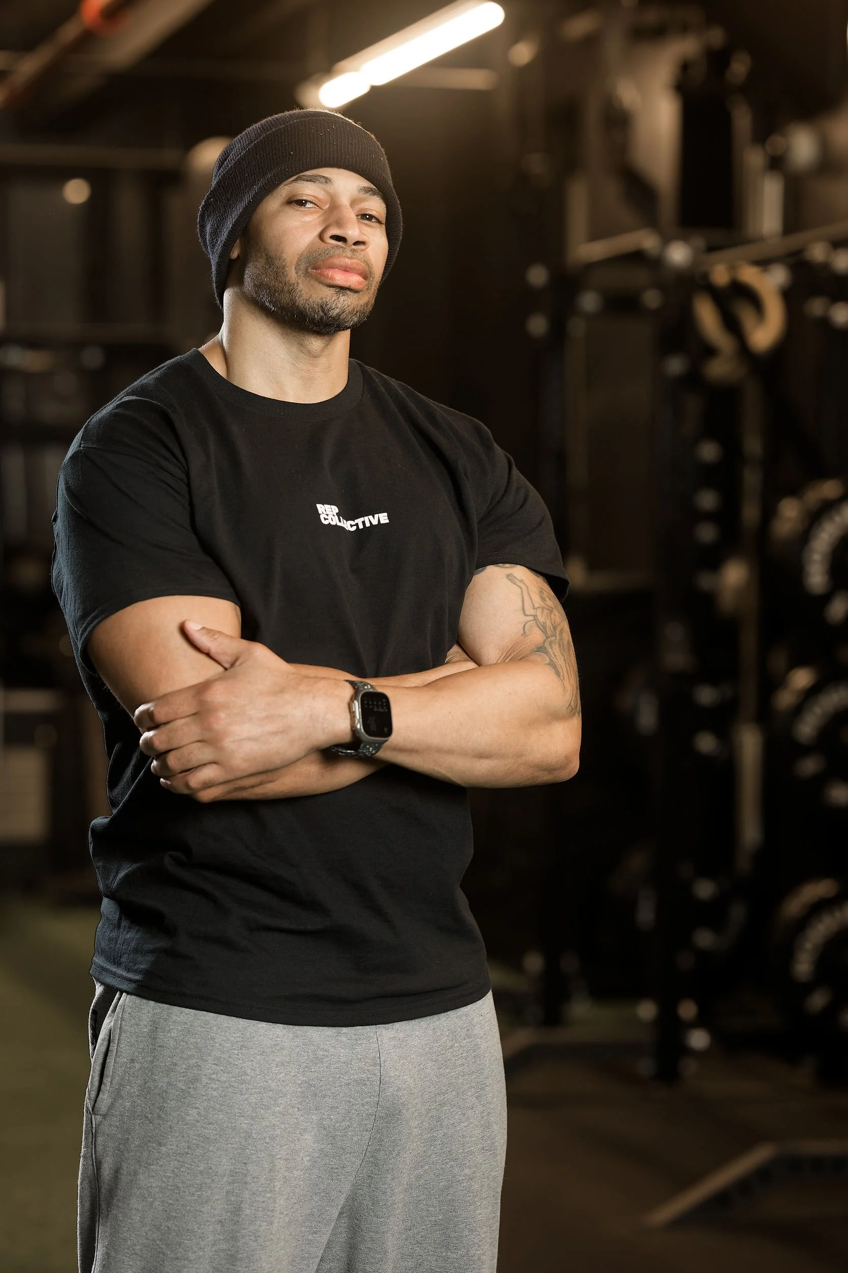 A muscular man wearing a black beanie, black T-shirt with 'REP COLLECTIVE' logo, and gray sweatpants standing with arms crossed in a gym with weightlifting equipment in the background.
