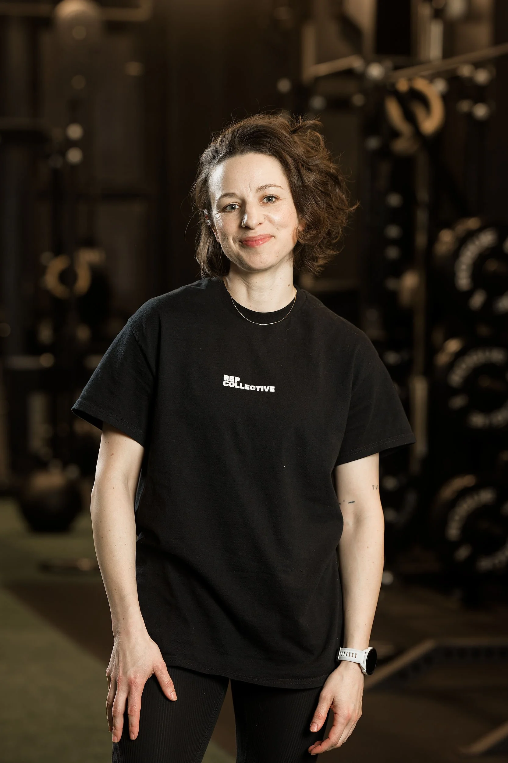 A woman with curly brown hair wearing a black t-shirt with white text, standing in a gym with weightlifting equipment in the background.