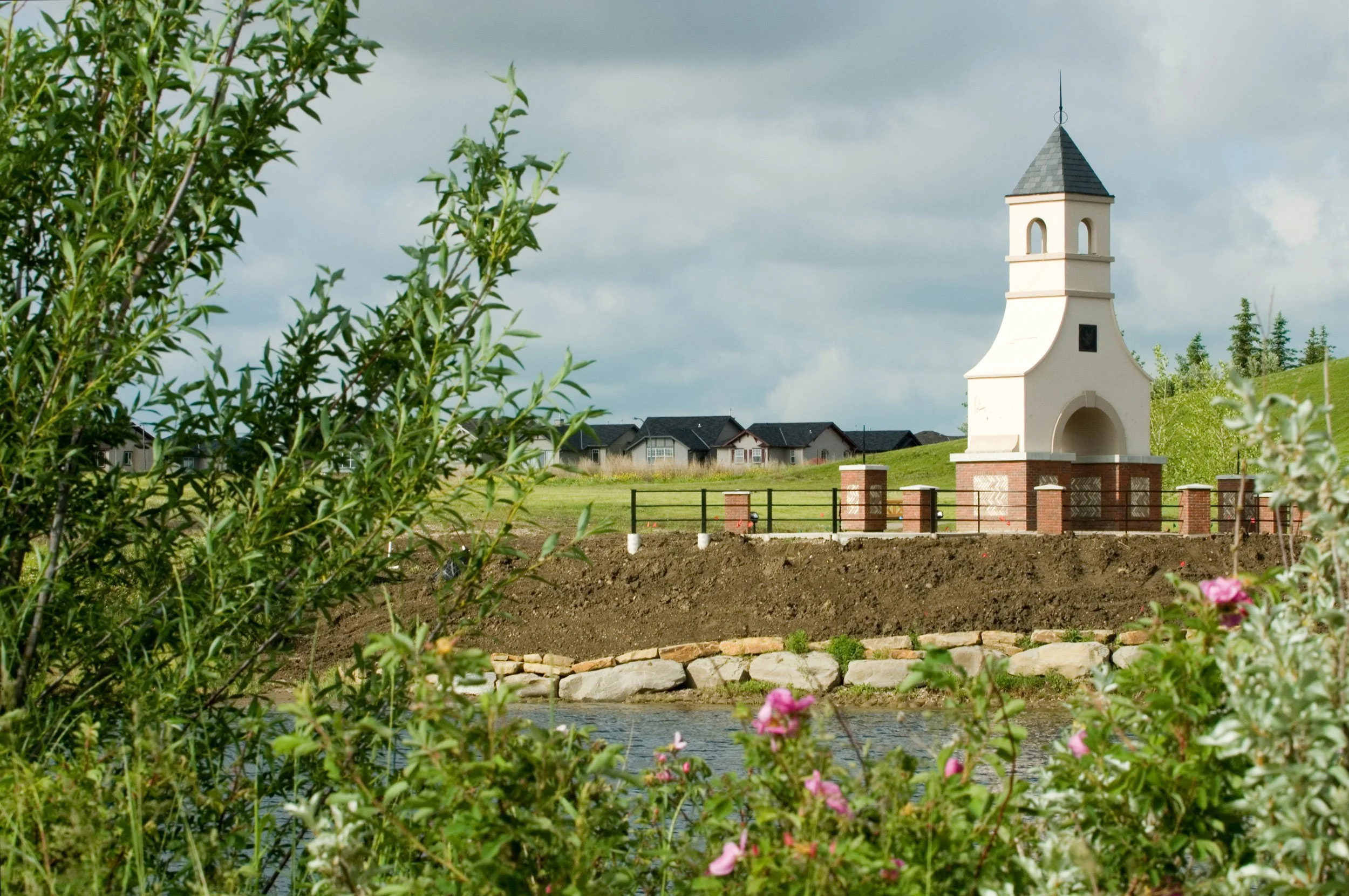 A small white chapel with a bell tower on a grassy hill, fenced with brick and metal, near a river, with houses in the background and a cloudy sky.