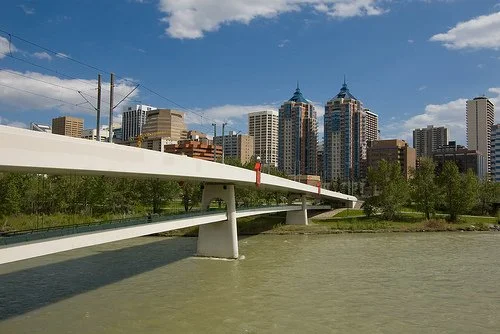 City skyline with high-rise buildings, a bridge over a river, trees on the riverbank, under a partly cloudy sky.
