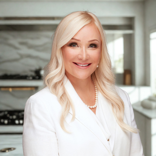 A smiling blonde woman wearing a white blazer and pearl necklace in a modern kitchen.