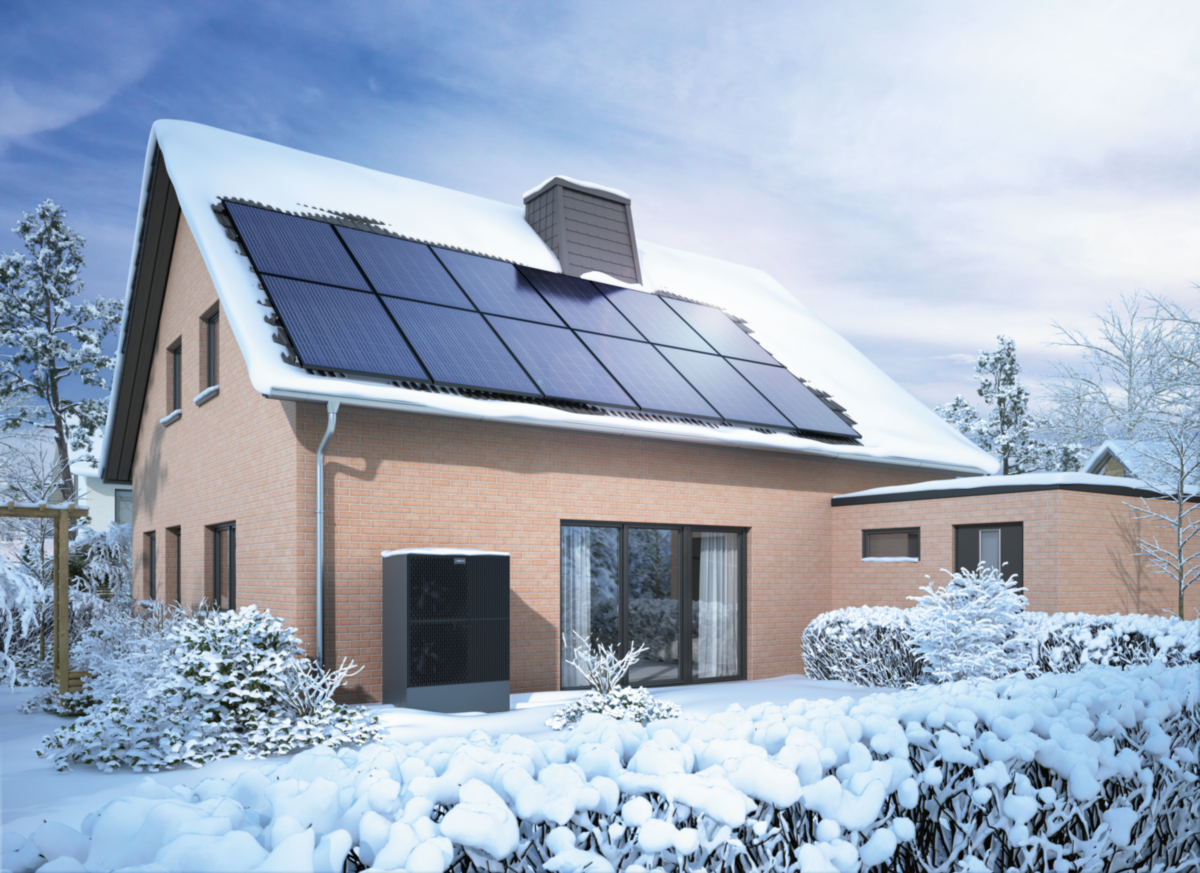 Ein Haus mit Solarpanelen auf dem schneebedeckten Dach, umgeben von verschneiten Büschen und Bäumen im Winter.