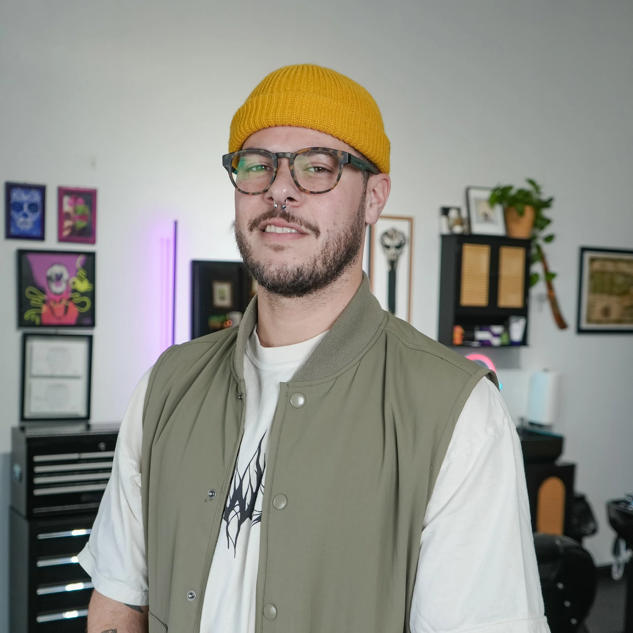 A young man with glasses, a beard, and a septum piercing, wearing a yellow beanie and a green vest over a white T-shirt, standing in a decorated room with framed art on the walls and a potted plant.