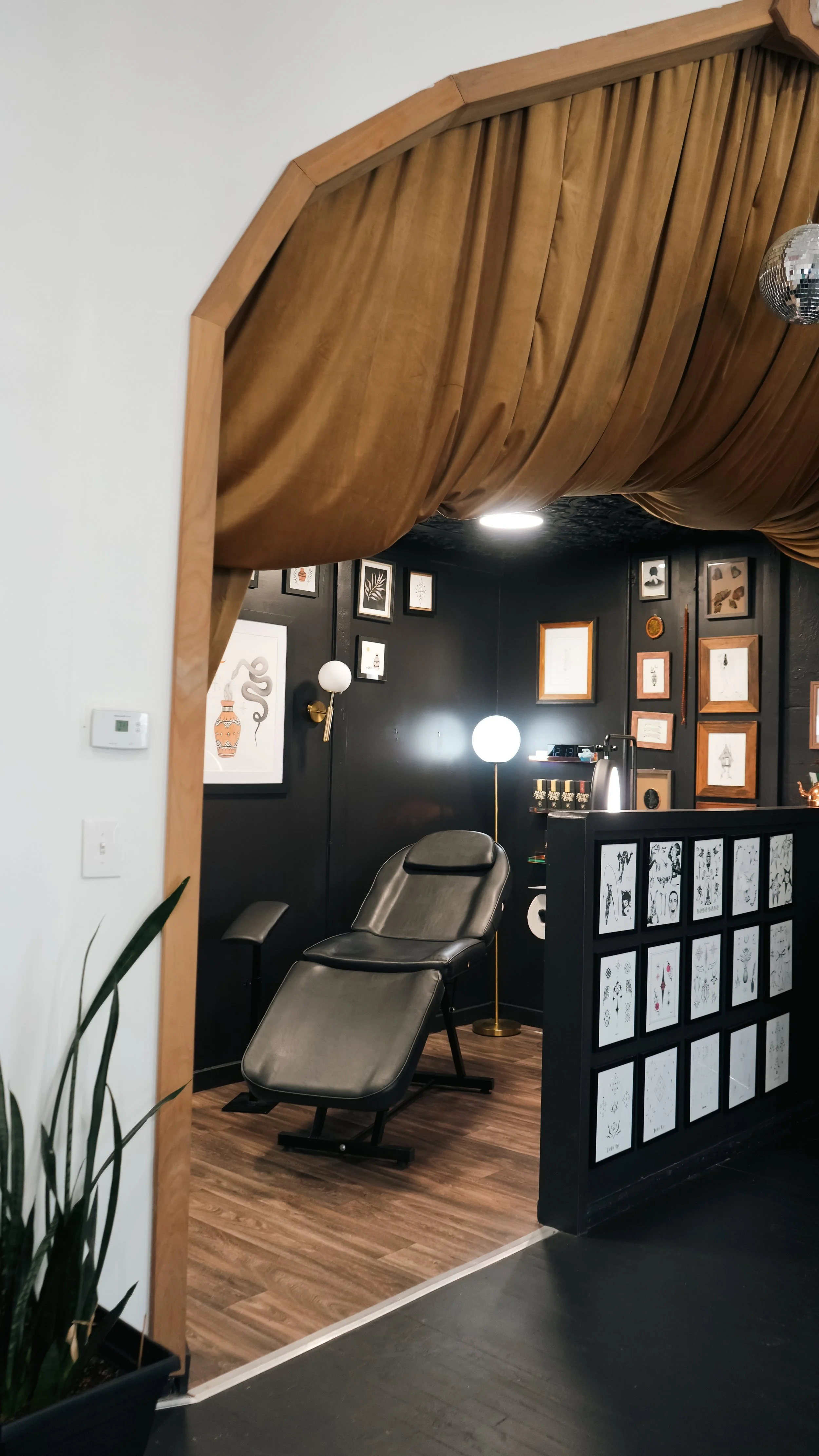 Interior room with a black wall decorated with framed art, a leather recliner chair, and modern lighting including a tall gold floor lamp with a globe light, viewed through a wooden-framed opening with a brown curtain.