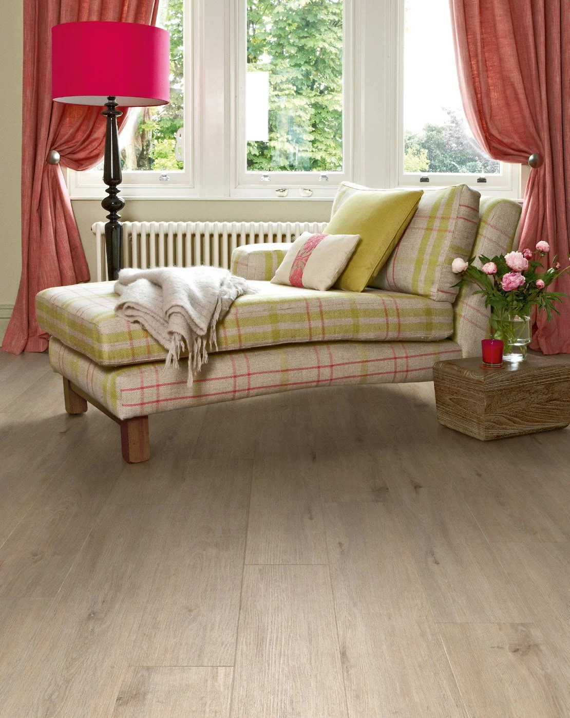 A cozy living room corner with a plaid upholstered chaise lounge, pink curtains, a pink table lamp, a glass vase of pink flowers, and a window view of trees outside.
