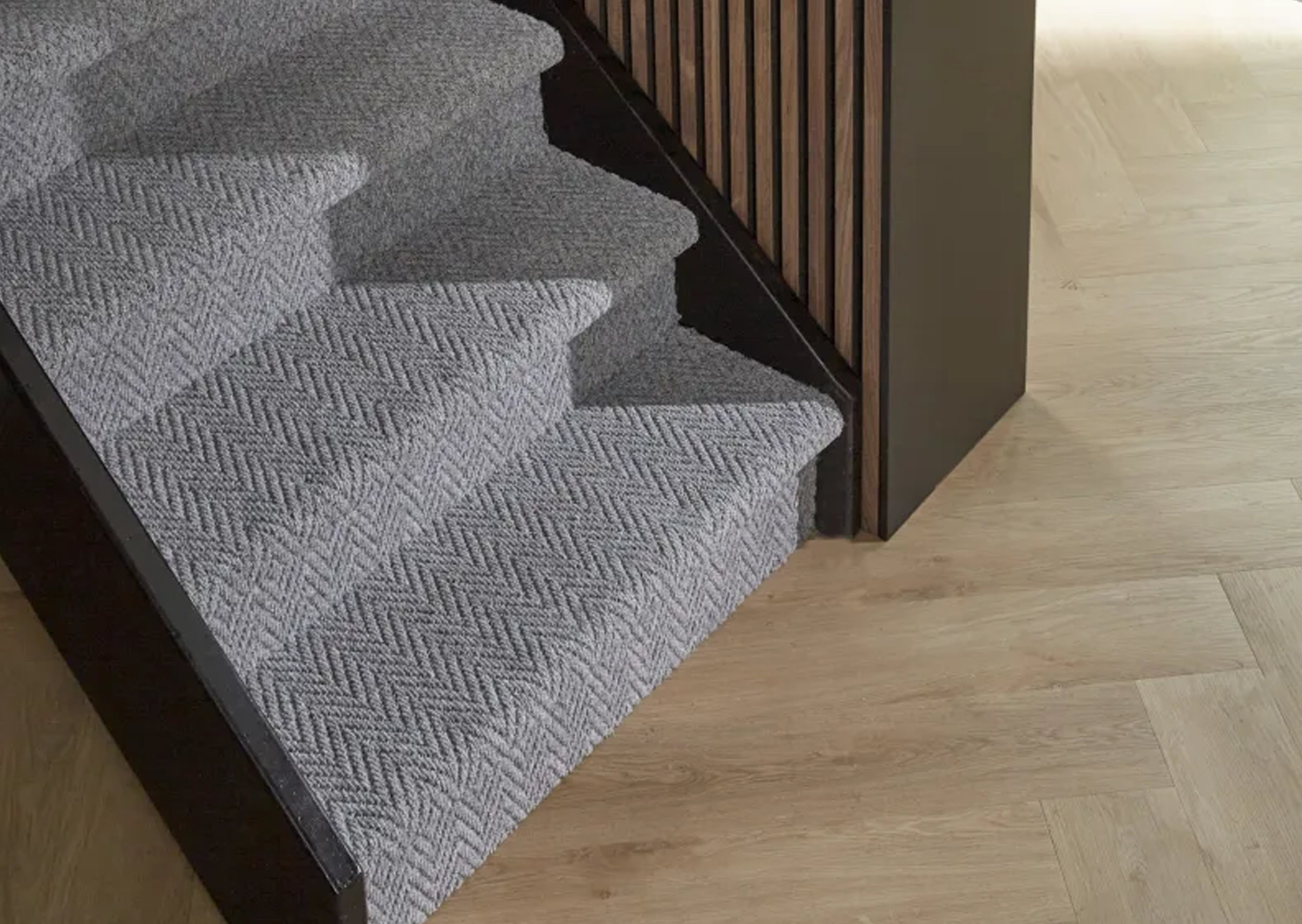 Close-up of a staircase with a grey textured carpet, dark wood side rail, and light wood flooring.