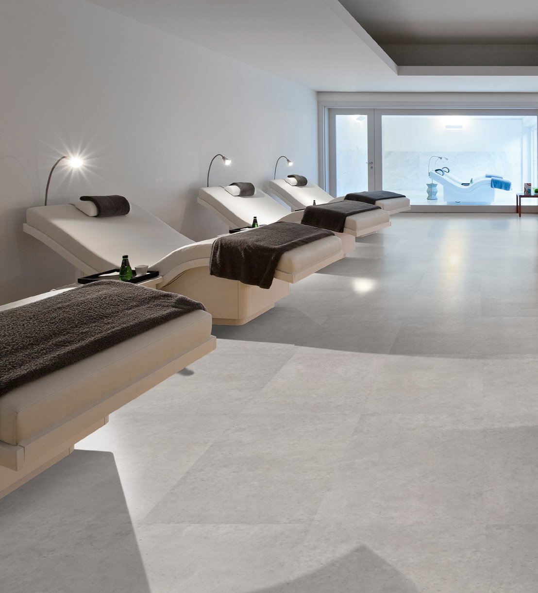 A minimalist spa room with four white lounge chairs lined up, each with a dark towel and small reading light. A large window outside reveals a hot tub on a terrace.